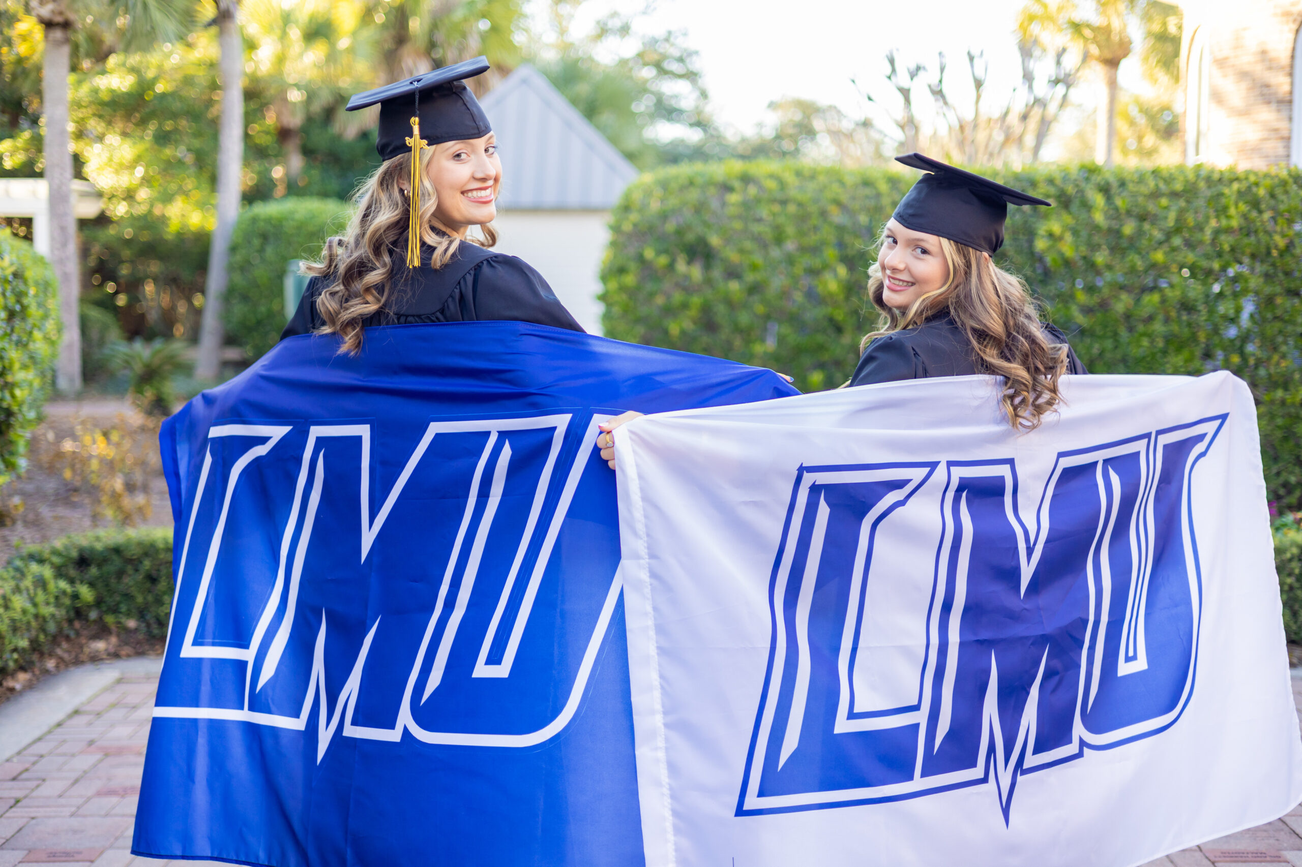 Two college graduates wearing black caps and gowns smile back toward the camera while holding large LMU flags behind them in a sunny outdoor setting with palm trees and greenery.