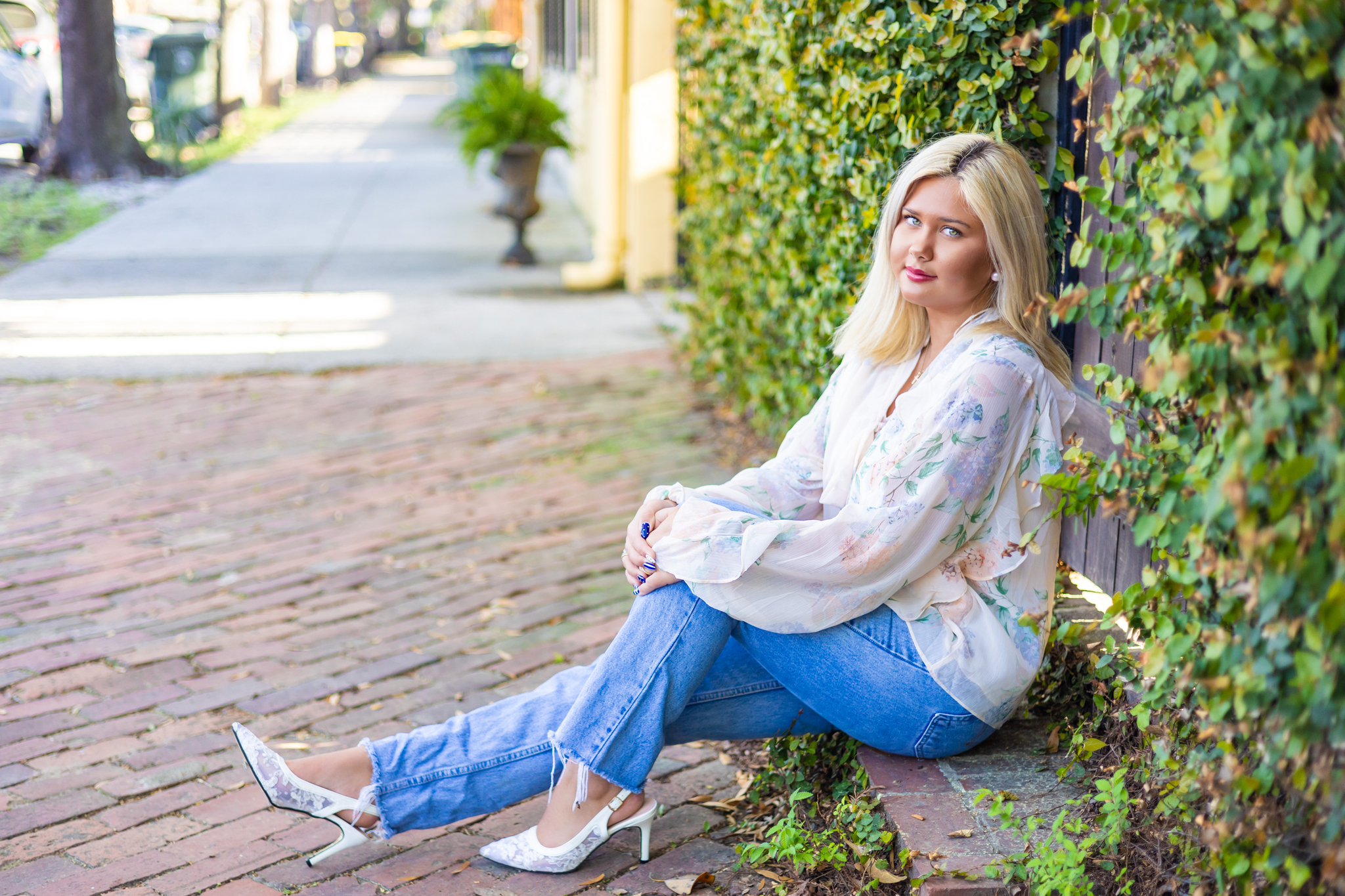 A high school senior sits against a vine covered fence in jeans and a floral blouse after finding great boutiques in Savannah
