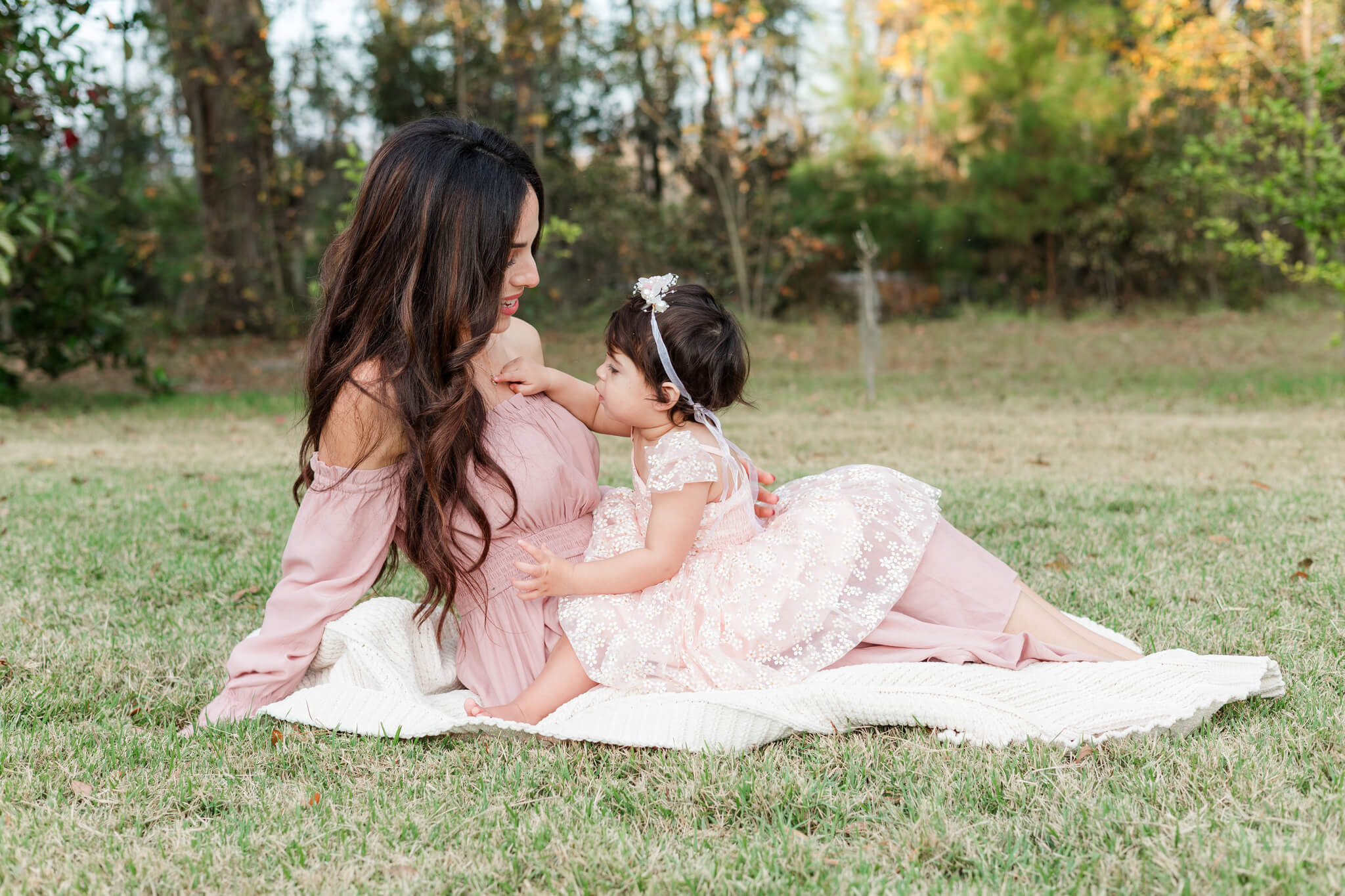 A toddler girl in a pink flower dress sits in mom's lap on a picnic blanket in a park