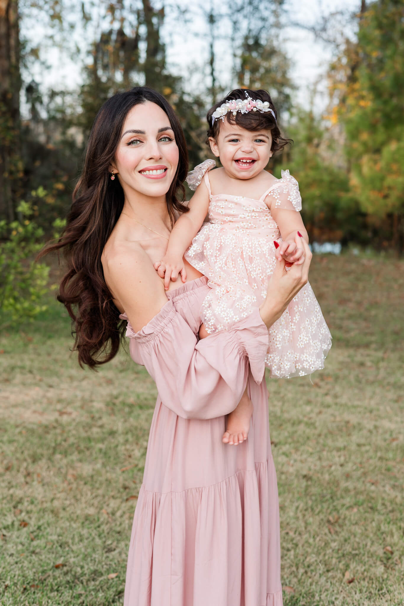 A smiling mother stands in a park holding her giggling toddler daughter in a pink gown after visiting indoor play space in Savannah