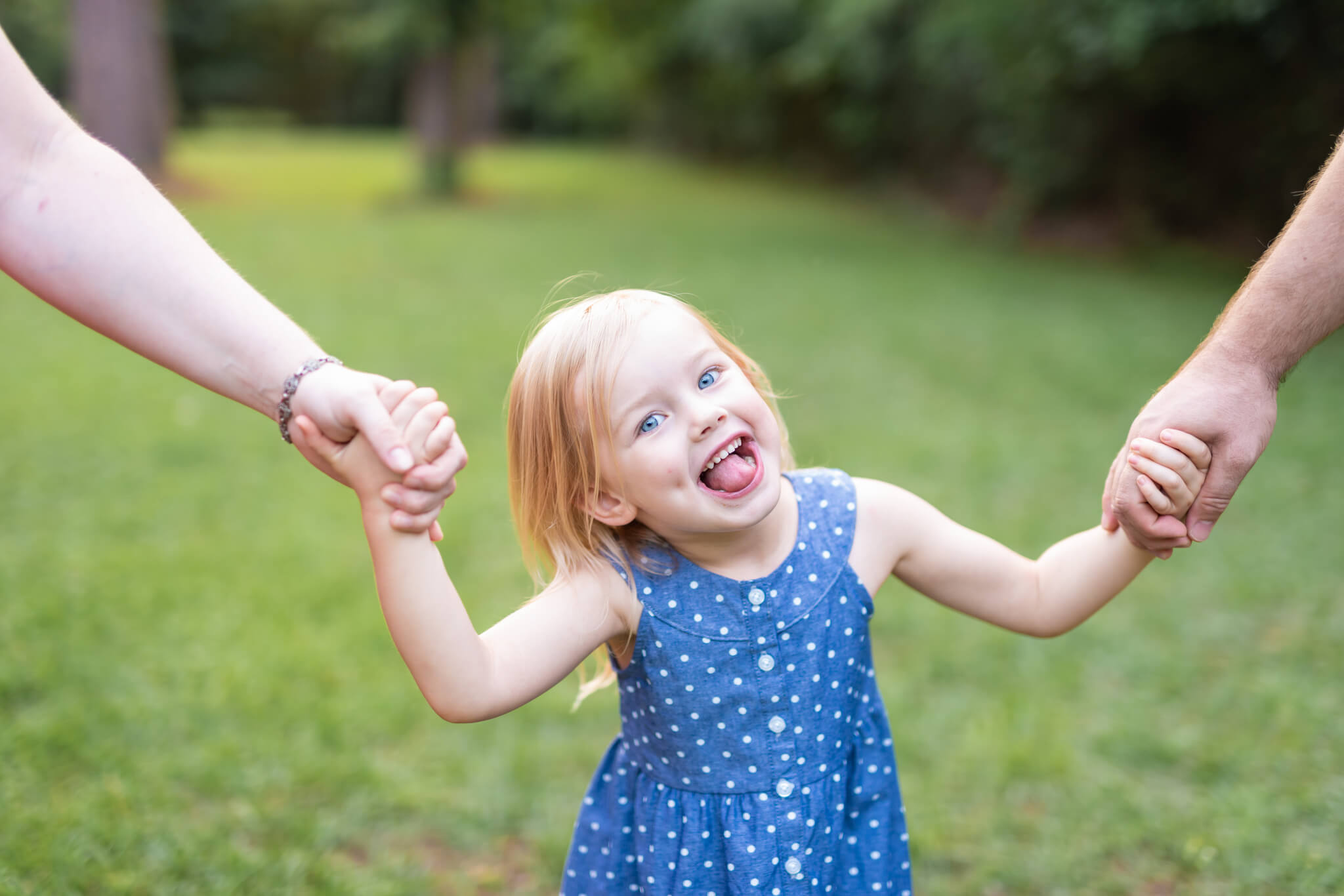 A toddler girl makes a silly face while holding mom and dad's hands in a park in a blue dress