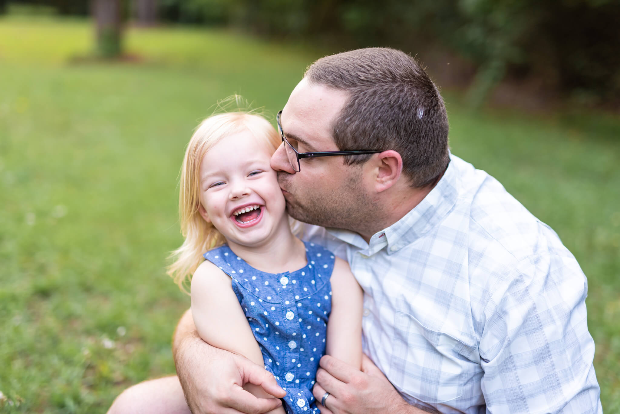 A laughing toddler girl in a blue dress is kissed by dad in a park lawn after visiting pediatricians in Savannah