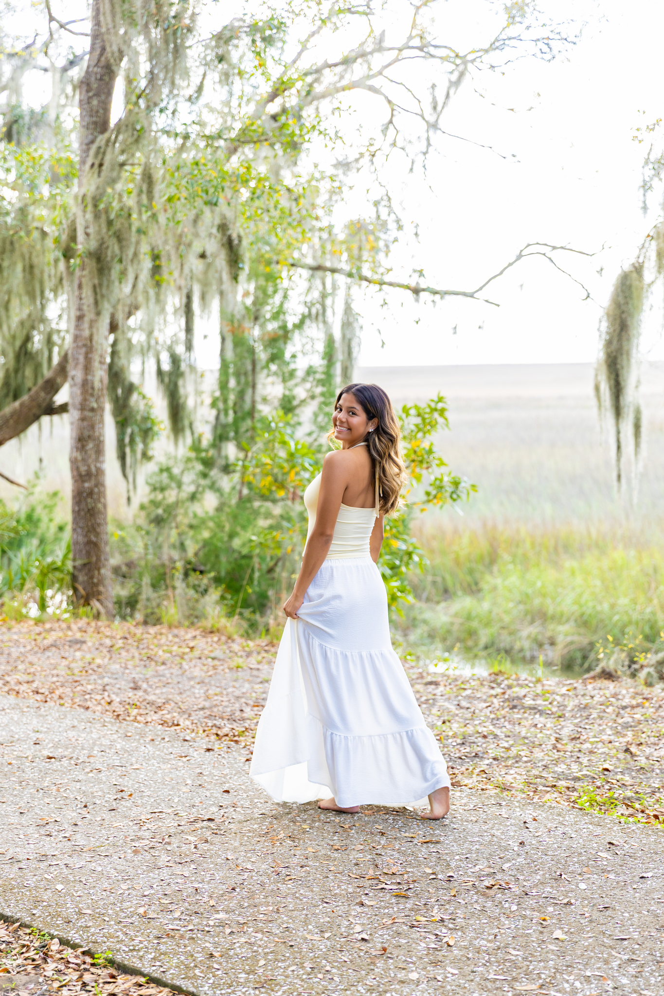 A smiling high school senior walks on a park sidewalk smiling over her shoulder in a white skirt after finding prom dresses in Savannah
