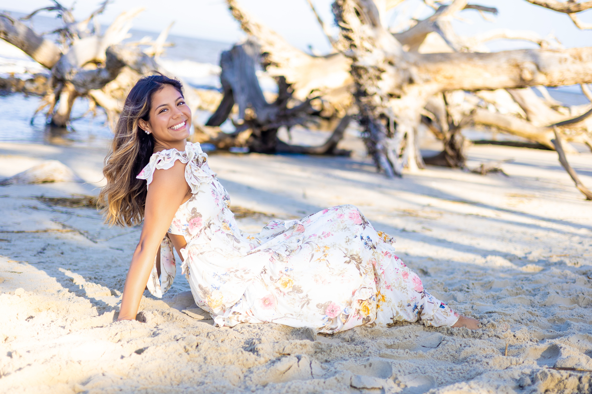 A happy high school senior sits in the sand on the beach in a floral print dress after shopping for prom dresses in Savannah