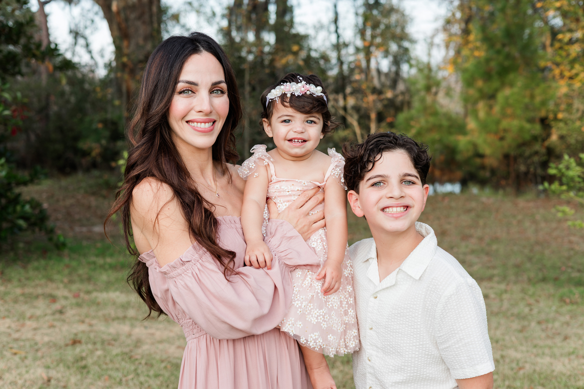 A happy mother in pink holds her young toddler daughter in a flower dress while smiling with her older son in a white shirt