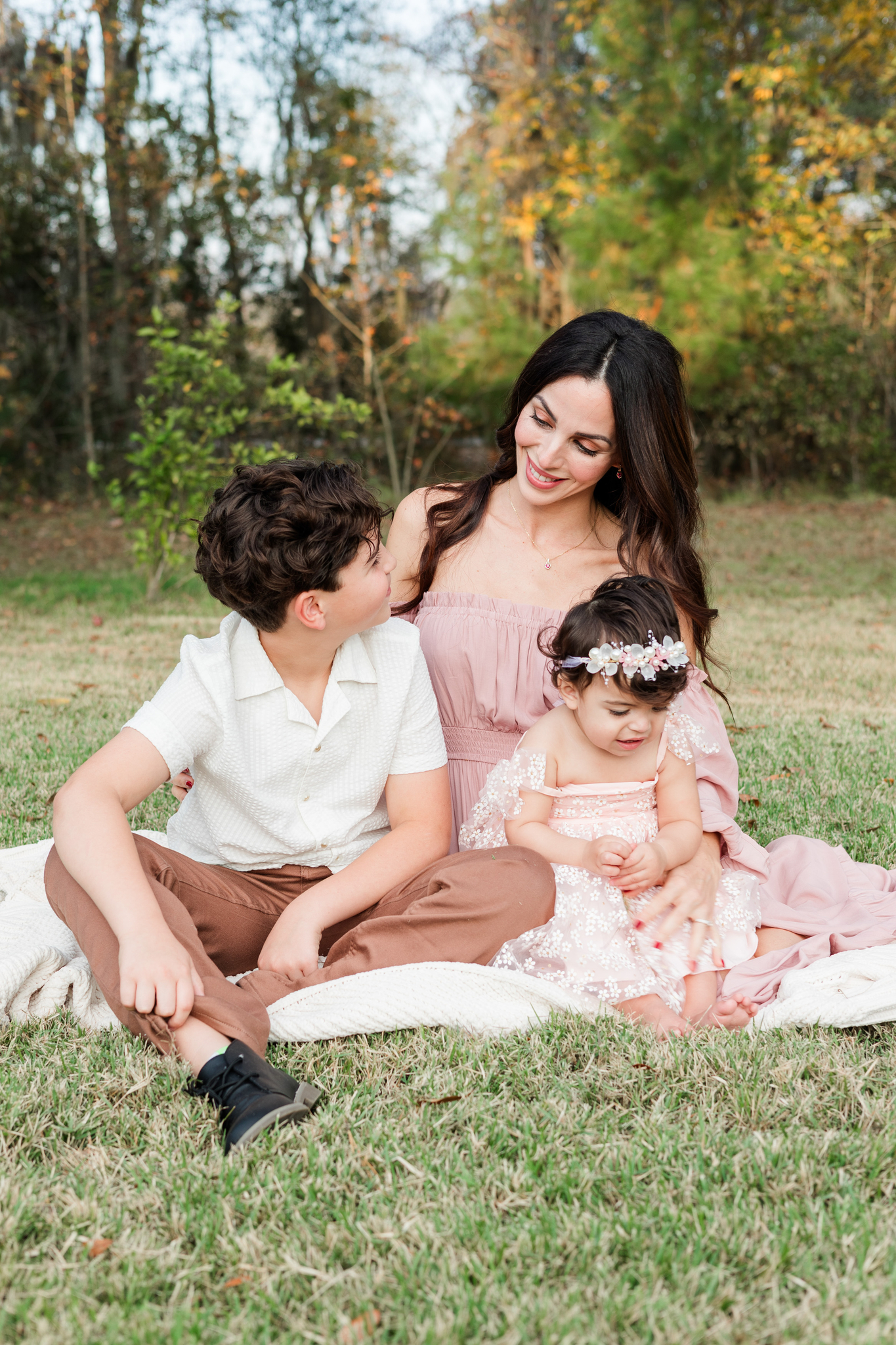 A happy mom in a pink dress sits in a park lawn with her toddler son and younger daughter on a blacnket before visiting toy stores in savannah