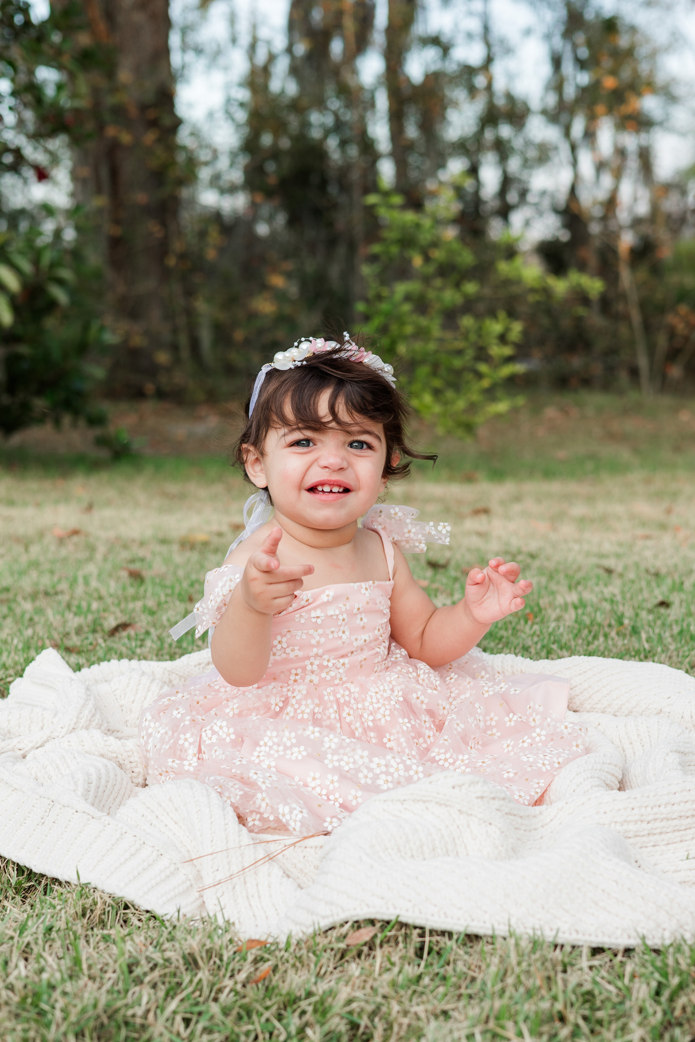 A toddler girl in a pink dress sits in a field on a picnic blanket smiling and pointing before visiting toy stores in savannah