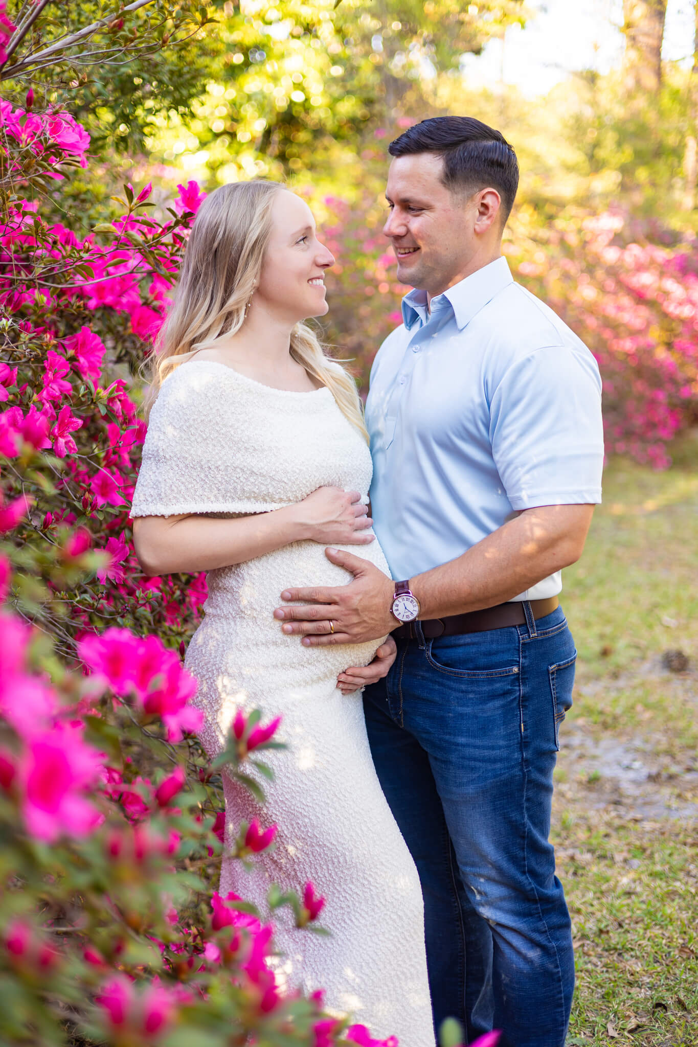 An expecting couple leans into a pink flower bush in a garden at sunset with hands on the bump