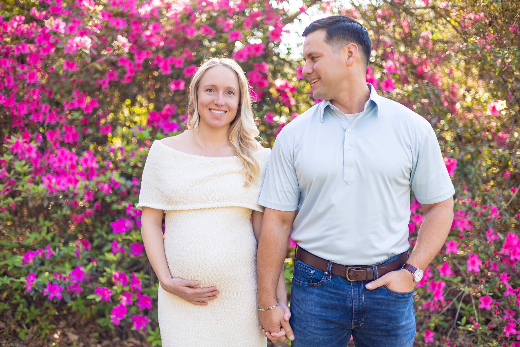 A mother to be in a white maternity gown stands in front of pink flower bush holding hands with her husband in blue after a couples massage in Savannah