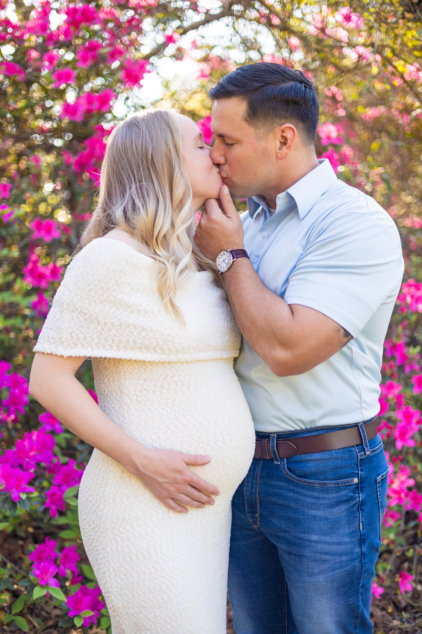 A happy pregnant couple in white and blue kiss in front of pink flowers at sunset after a couples massage in Savannah