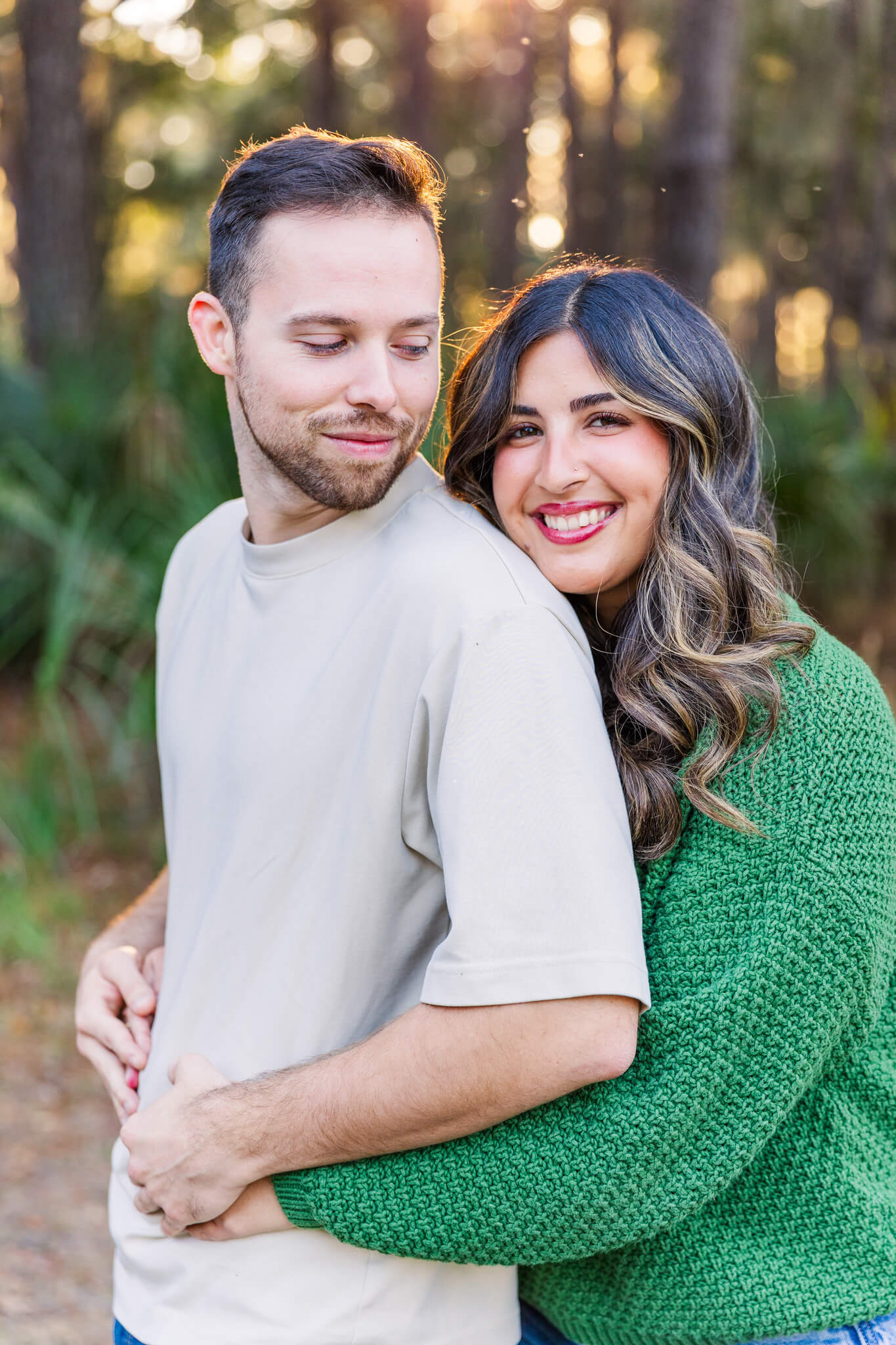 A woman in a green sweater hugs her fiancee from behind in a park at sunset as they both smile after enjoying date ideas in savannah