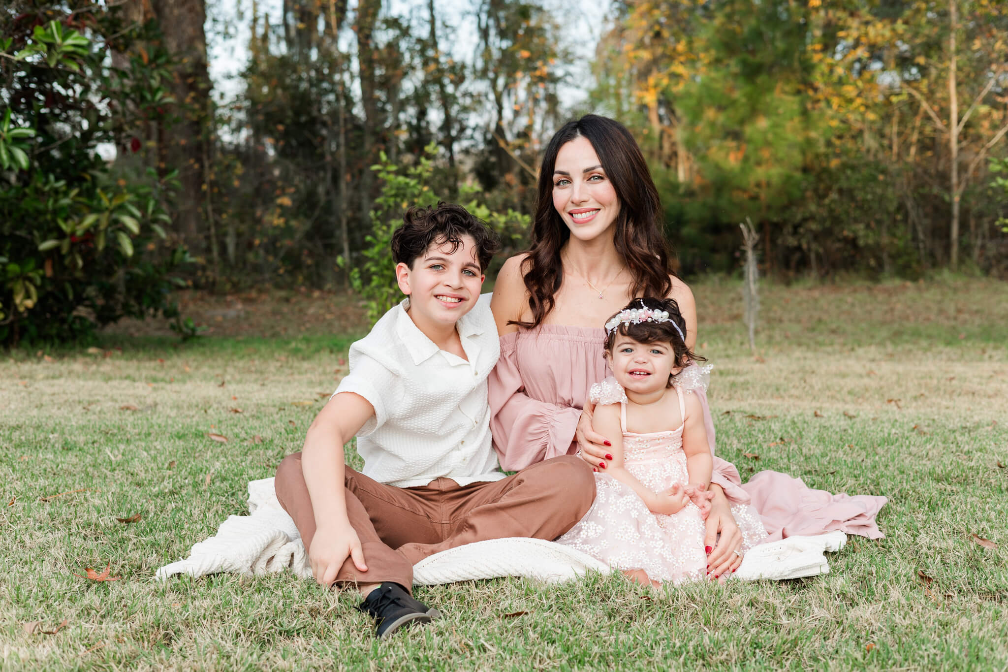 A happy mom smiles while sitting in a park lawn at sunset with her toddler daughter and young son