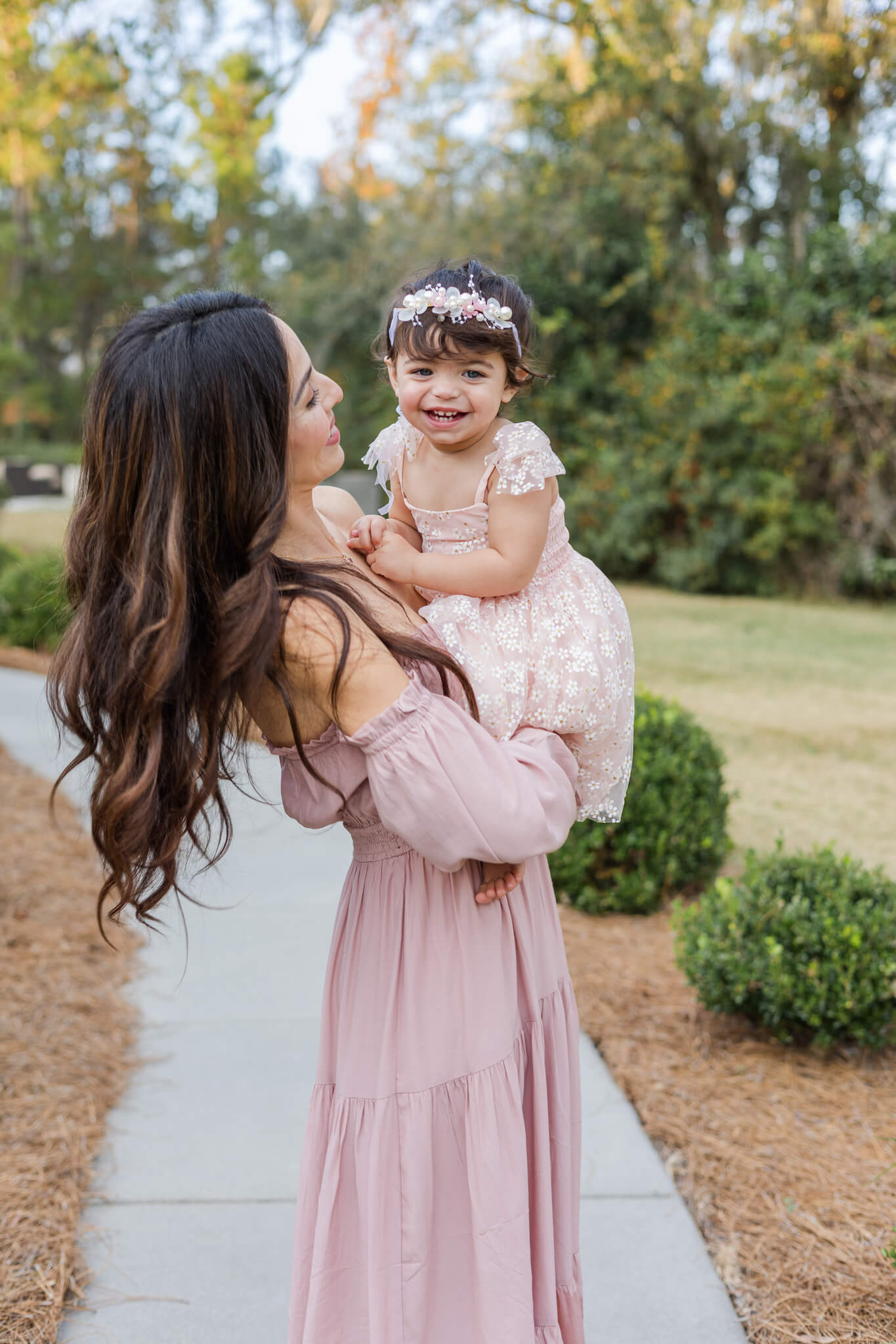 A giggling toddler girl sits in mom's arms in a park in pink dresses after exploring daycares in Savannah