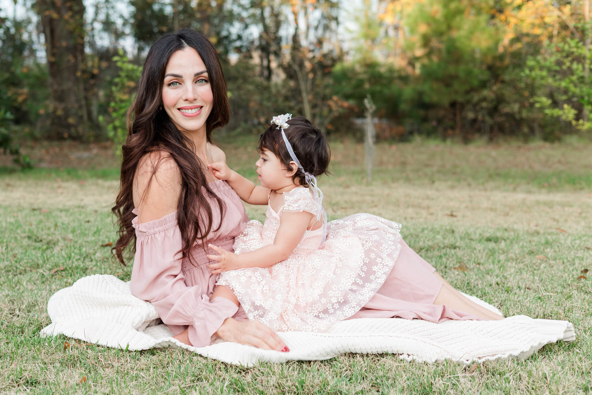 A happy mom sits on a picnic blanket in a park with her toddler daughter playing in her lap after visiting daycares in Savannah
