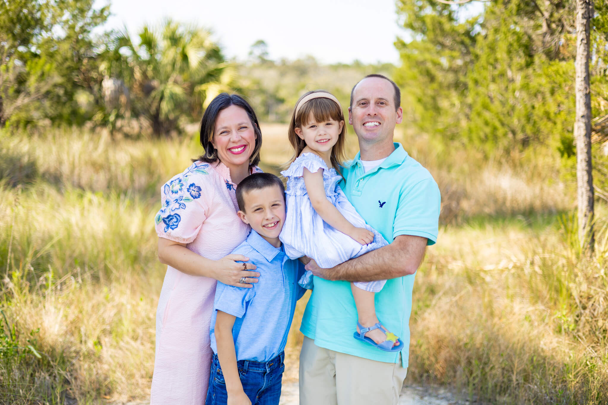 A mom and dad stand in a park trail with their toddler son and daughter in bright spring colors