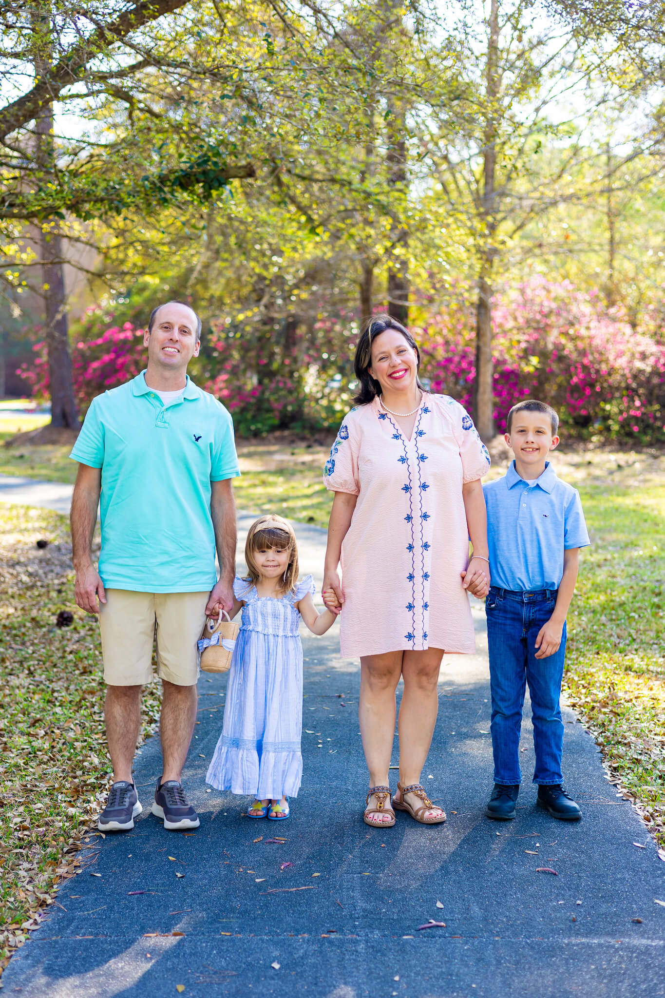 A mom and dad stand in a park sidewalk holding hands with their toddler son and daughter after finding a nanny in savannah ga