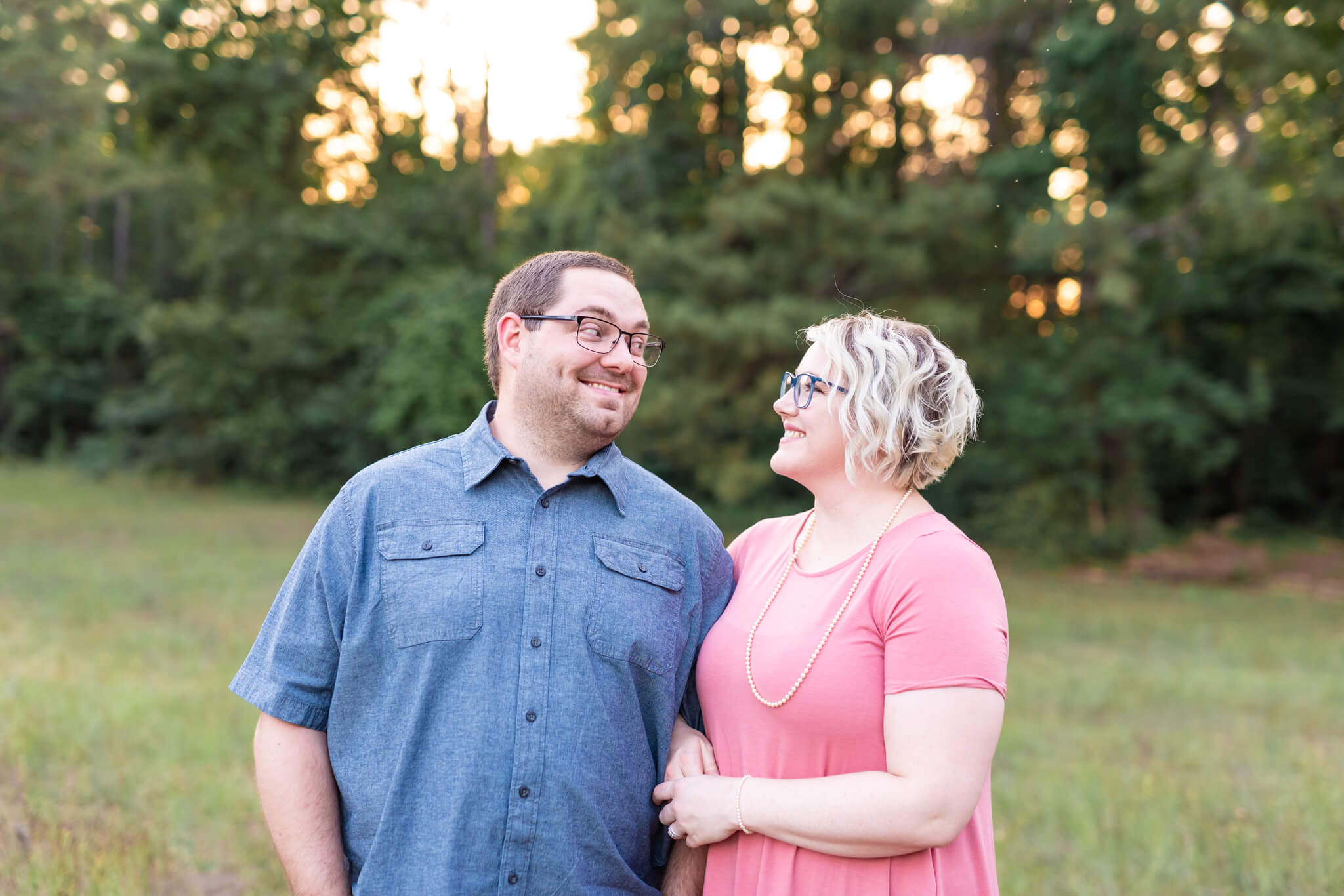 A happy man smiles big at his wife in pink while standing in a park field at sunset