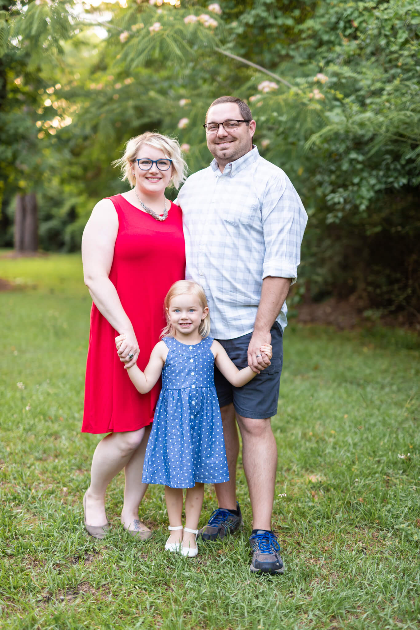 Happy mom and dad in red and white stand holding hands with toddler daughter in a park in her blue dress after finding preschools in savannah ga