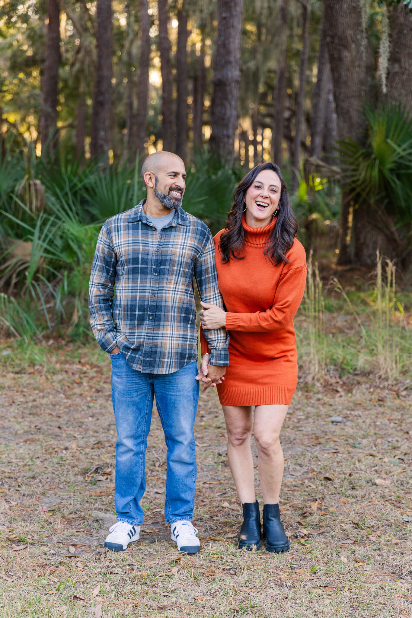 A woman laughs while standing holding hands with her husband in a park at sunset in a red dress and plaid shirt after visiting romantic restaurants in savannah