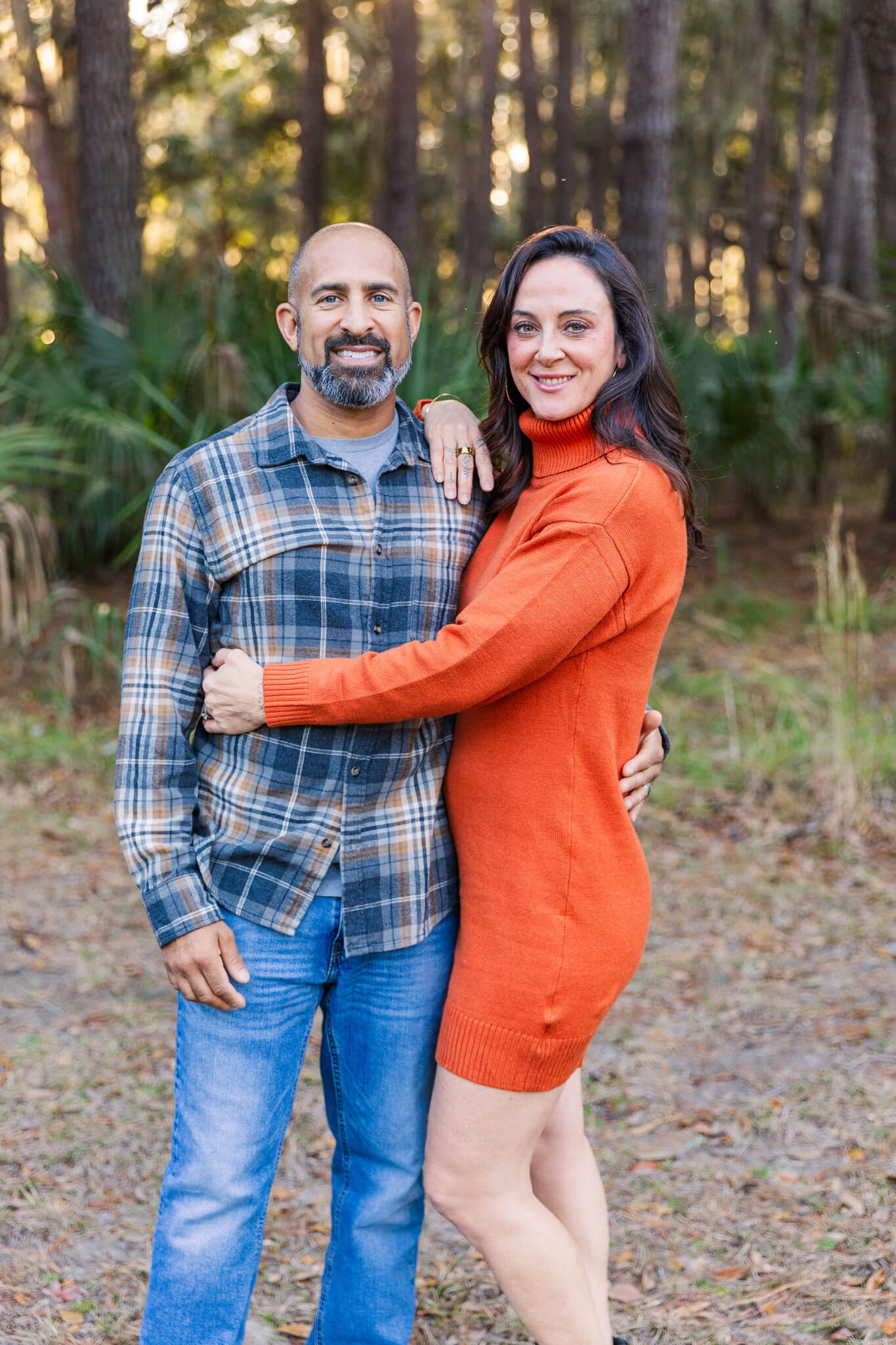 A happy mom and dad in orange and plaid hug and smile while standing in a park at sunset after visiting romantic restaurants in savannah