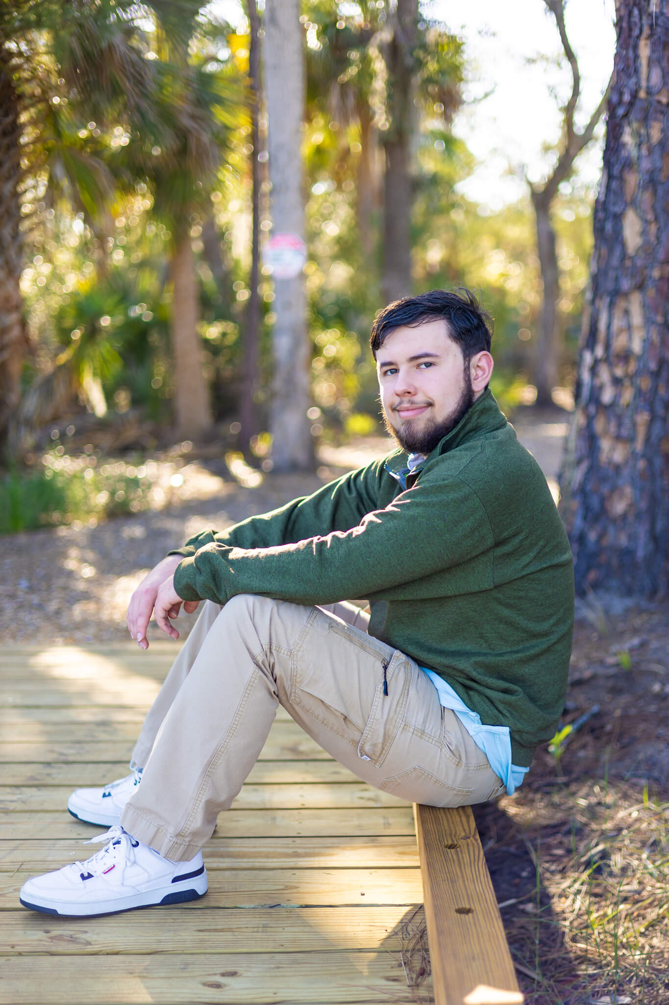 A teenager sits on a wooden boardwalk at sunset smiling leaning on his knees in a green sweater