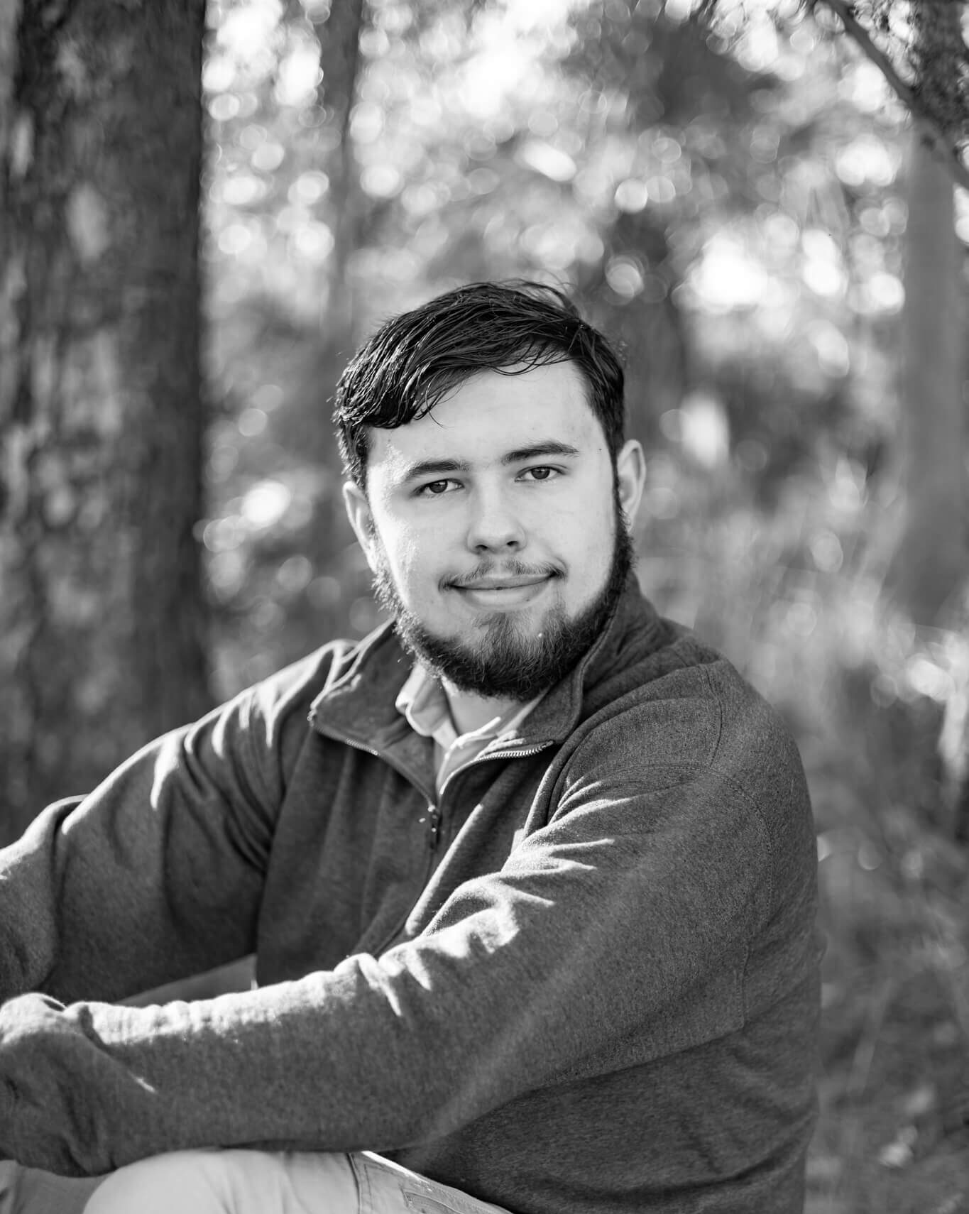 A black and white image of a high school grad sitting in a forest at sunset in a sweater after finding tux rental in Savannah, GA