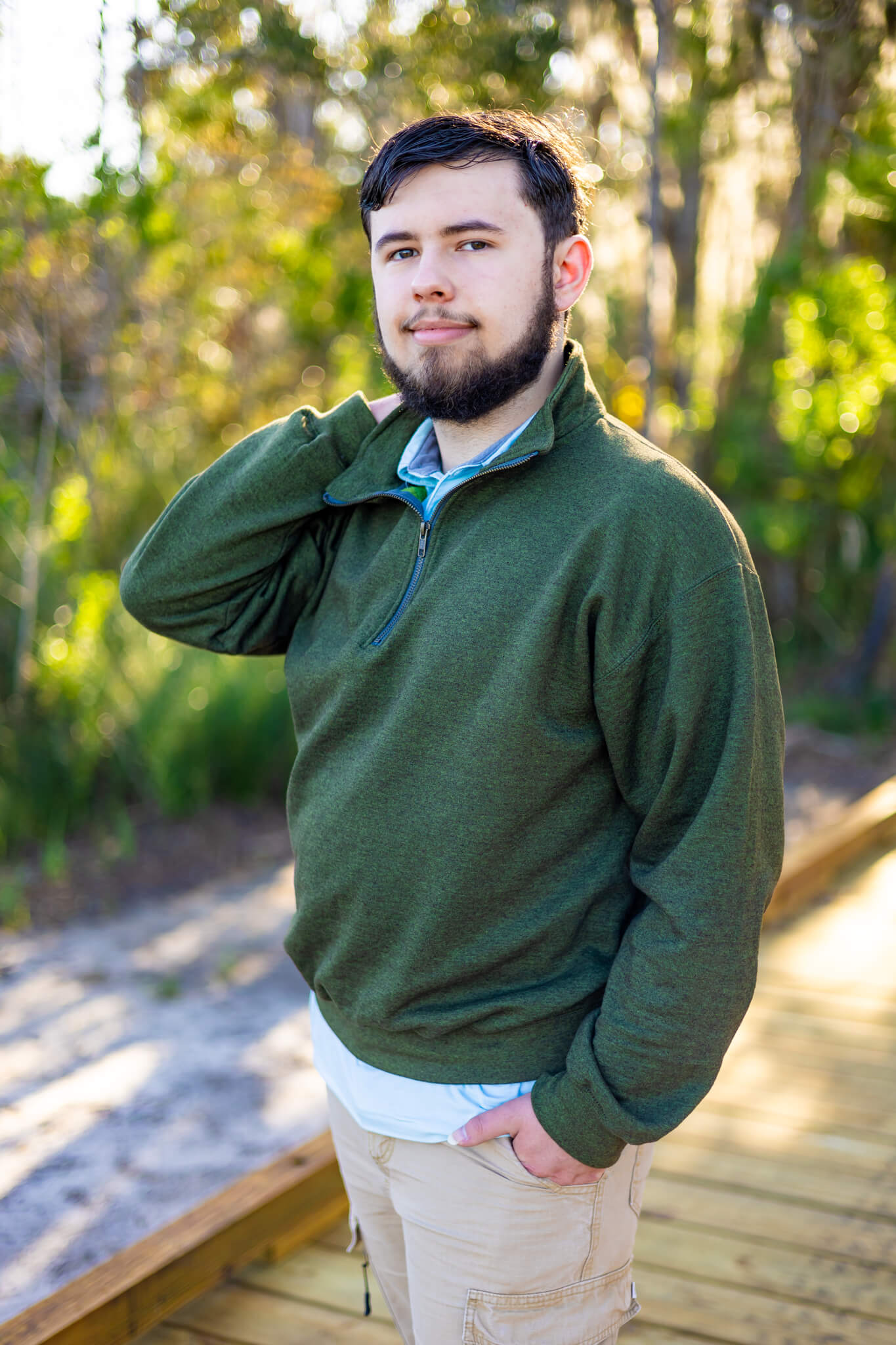 A high school senior in a green sweater walks on a beach boardwalk at sunset after finding tux rental in Savannah, GA