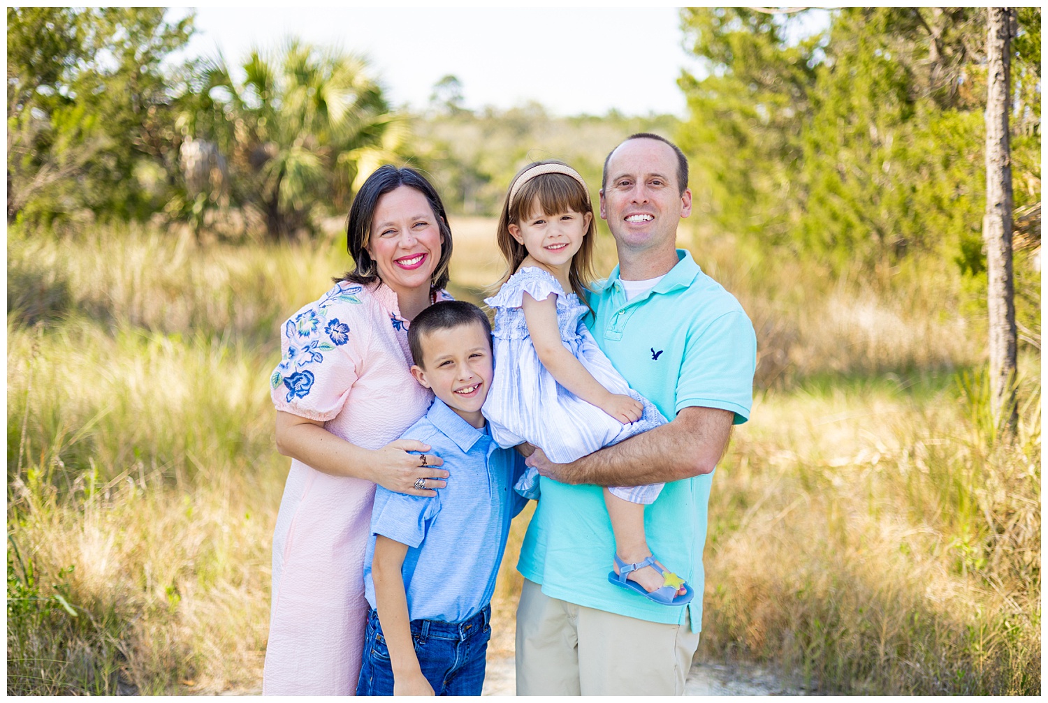Savannah family of four posing outdoors on a sunlit sandy path, with parents smiling and holding their young daughter while their son stands in front, surrounded by soft greenery and palm trees.