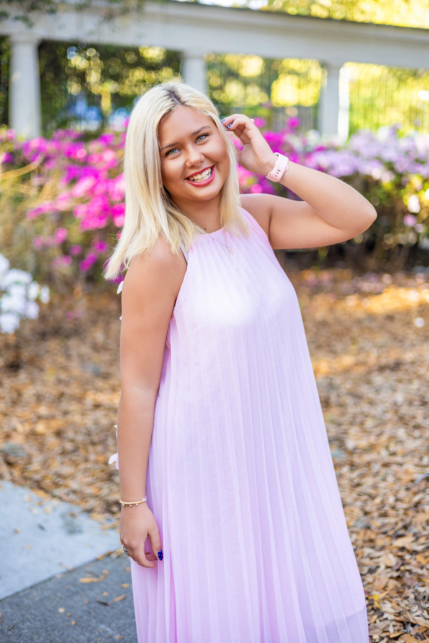 A high school senior in a pink gown smiles while pulling hair behind her ears in a garden with colorful azaleas after visiting hair salons in savannah ga