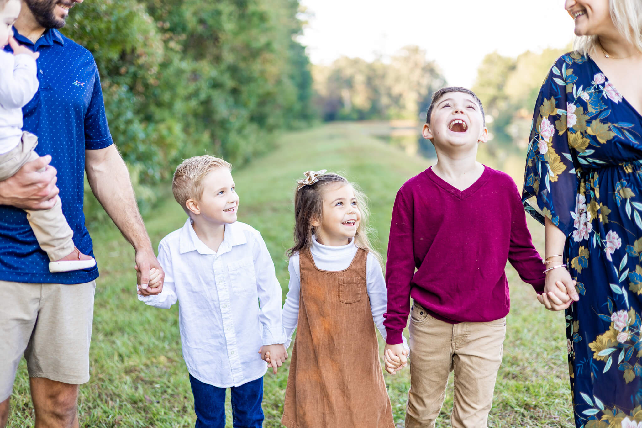 Three happy siblings hold hands with mom and dad while walking in a park by a pond with big smiles and laughs thanks to marriage counseling in savannah