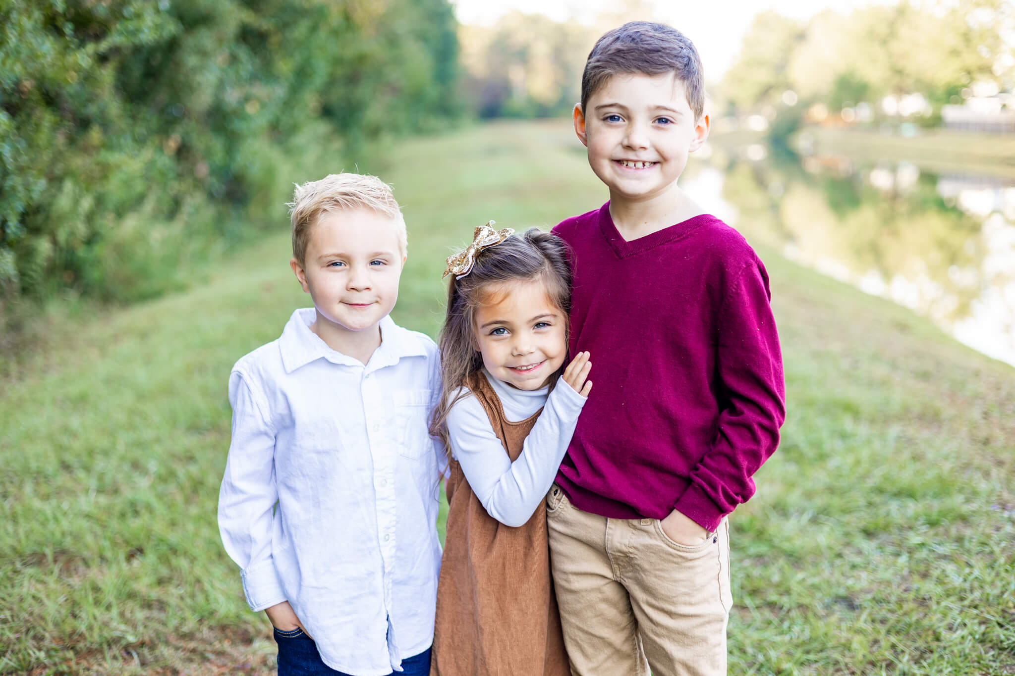 Three young siblings stand smiling by a pond hugging each other