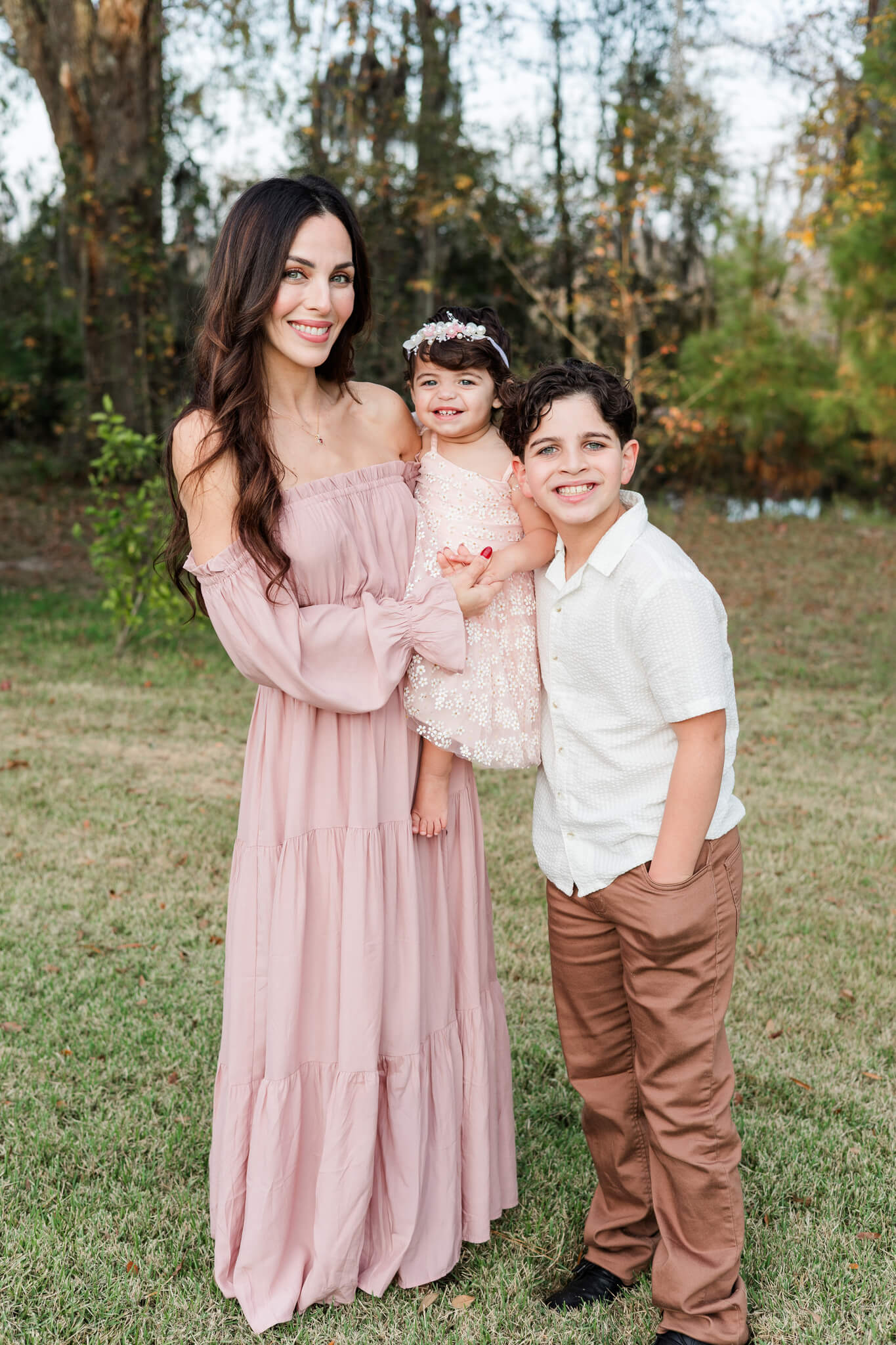 A young boy stands smiling in a white shirt and hugging his baby sister and mom in a park lawn