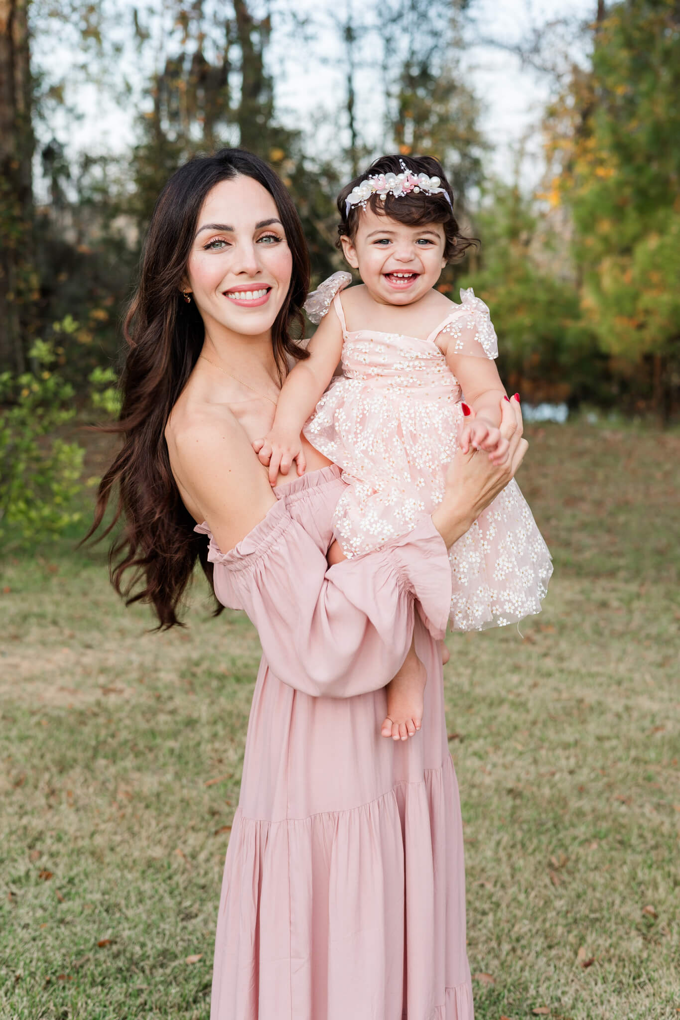 A giggling baby girl in a pink dress sits in mom's arms while she stands in a park after discovering montessori schools in savannah