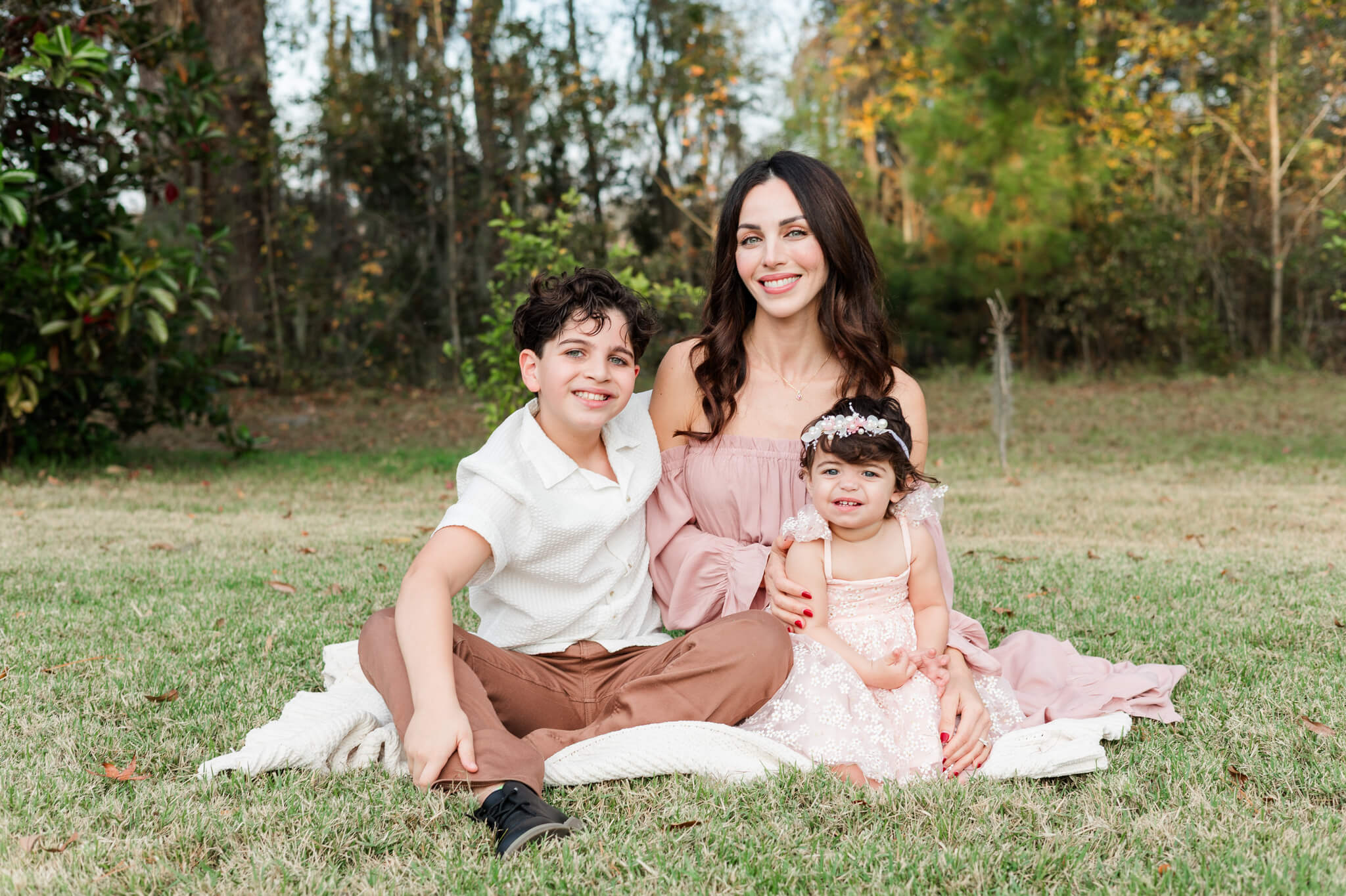 A happy mother in a pink dress sits in a park lawn on a picnic blanket with her young son and toddler daughter after exploring montessori schools in savannah