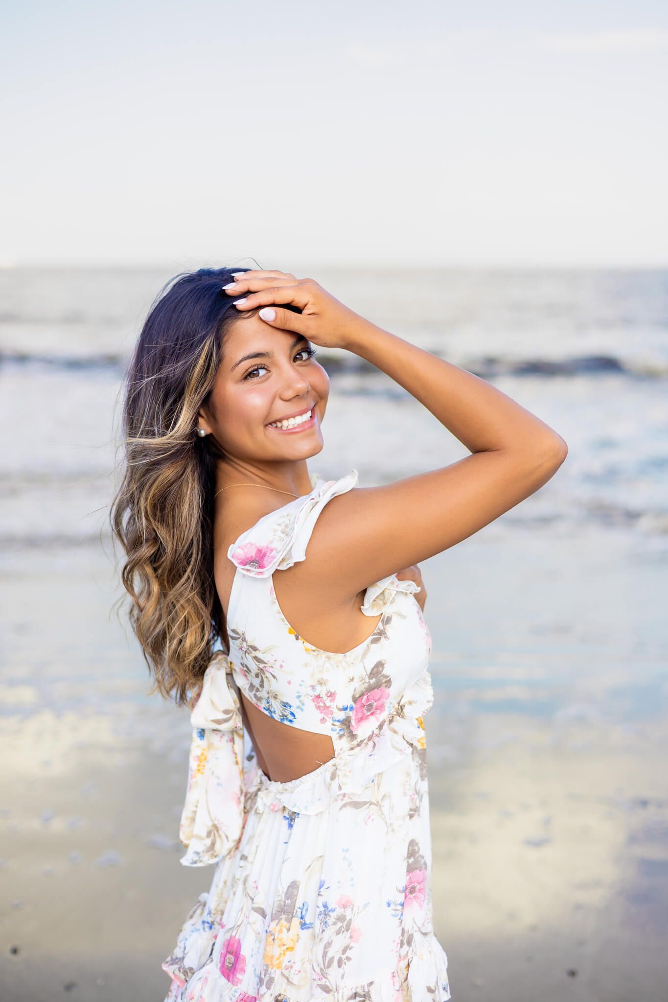 A smiling high school grad with a hand in her hair smiling over her shoulder on a beach