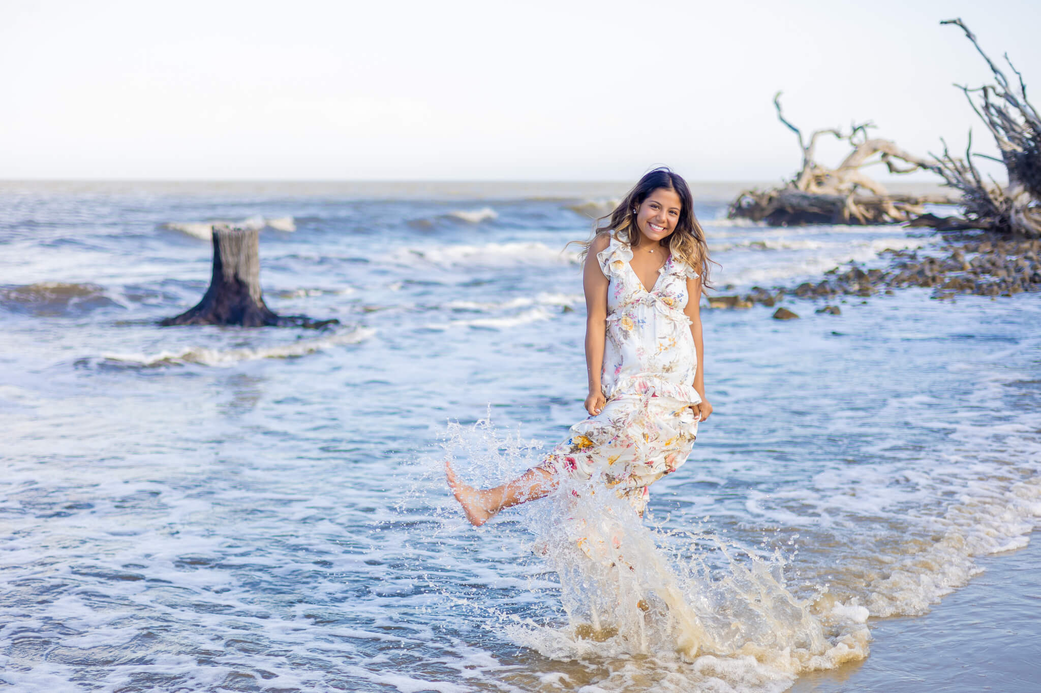 A happy high school senior kicks the water on the beach at sunset in a white flower print dress after finding great nail salons in savannah ga