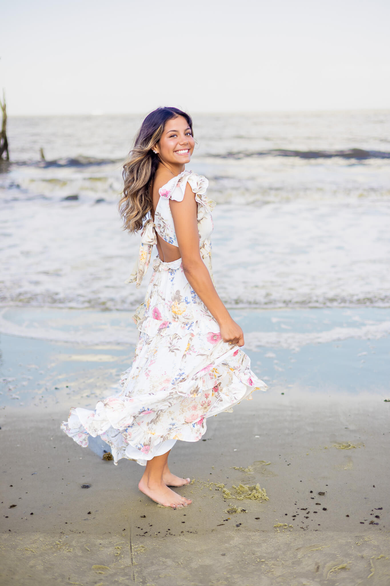 A smiling high school senior in a flower print dress twirls on the beach while looking over her shoulder after finding nail salons in savannah ga