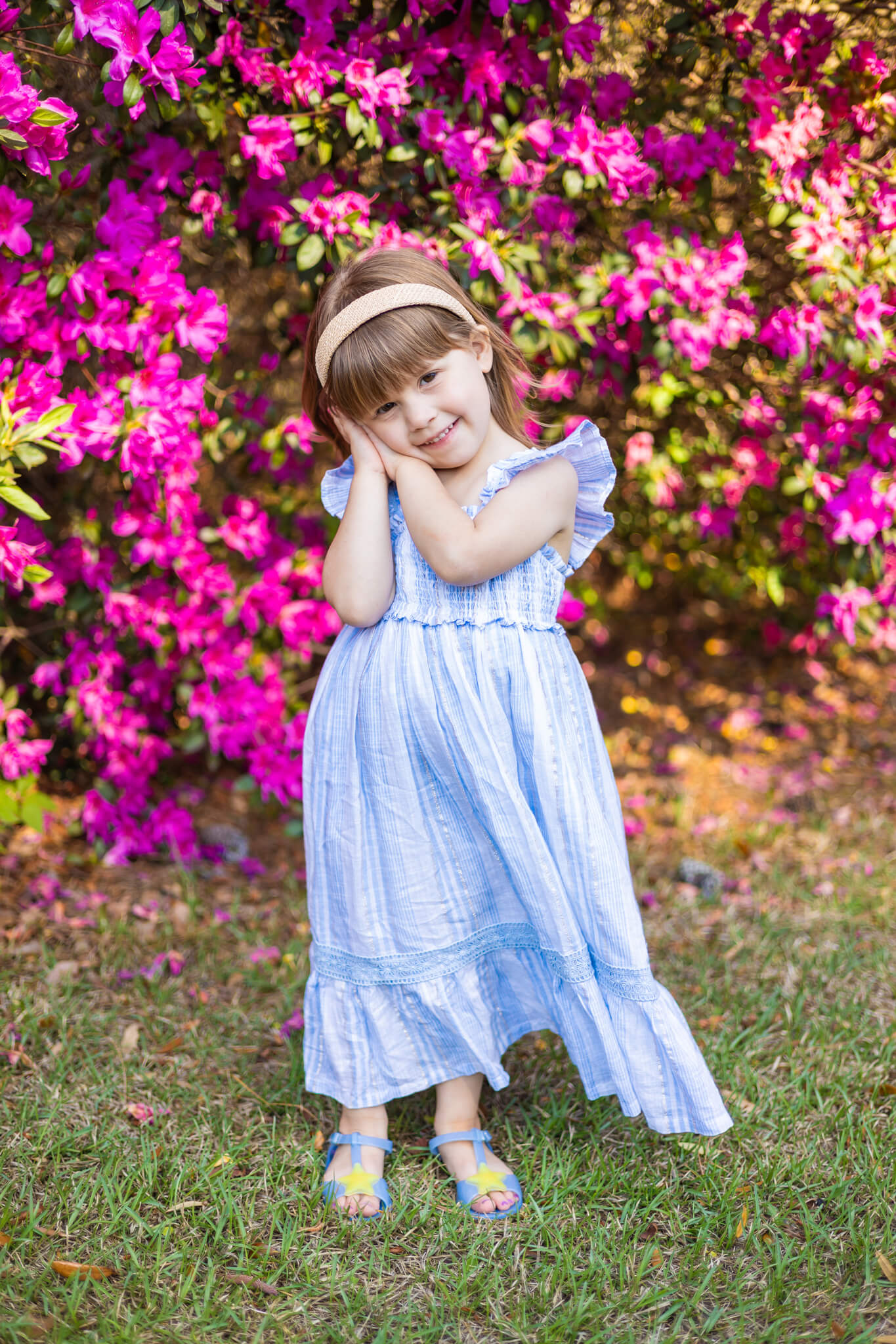 A toddler girl smiles with hands on her cheek in a blue dress in front of pink azaleas