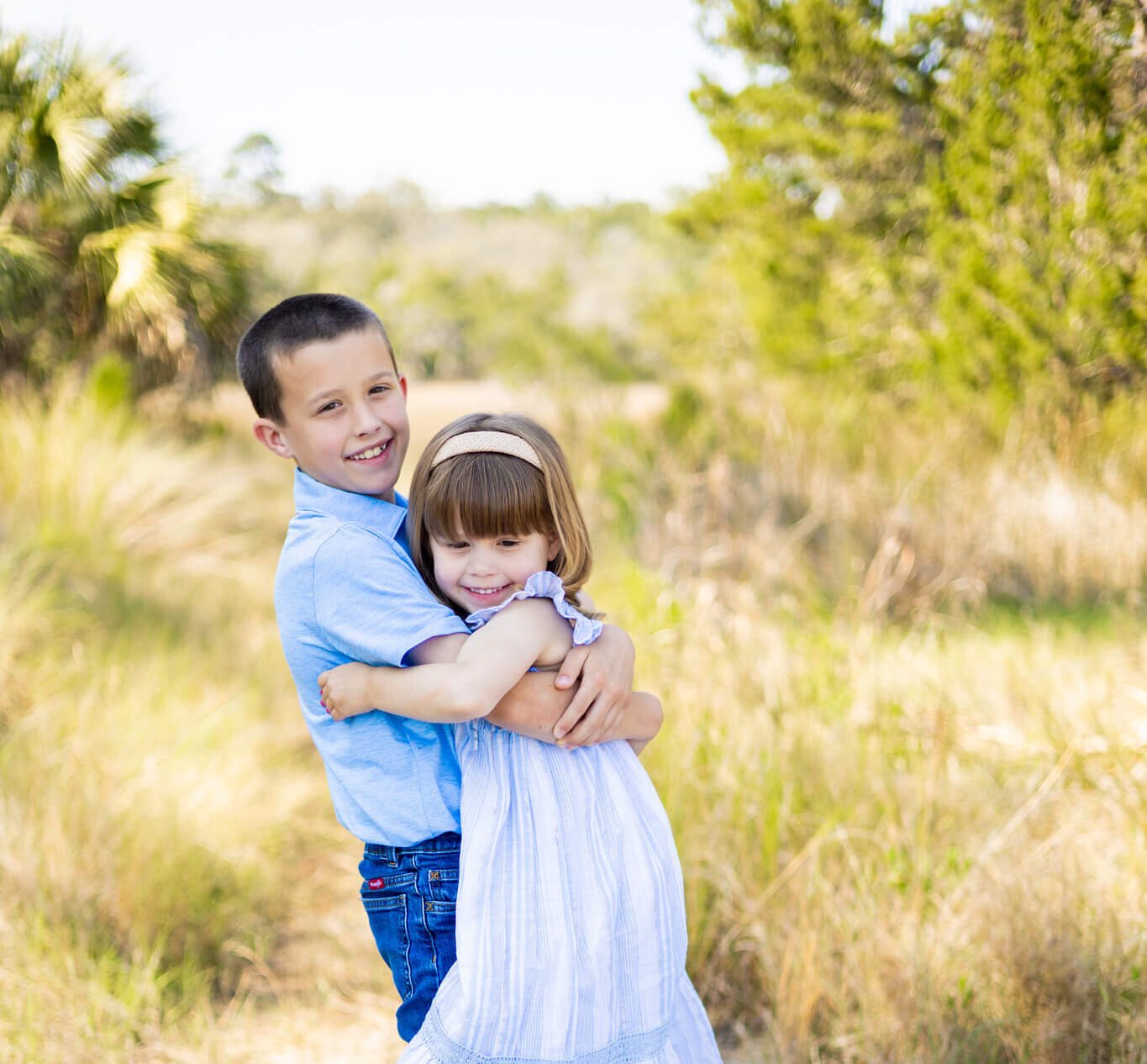 A toddler girl is hugged and lifted by bigger brother in blue dress and blue shirt in a park while smiling after visiting pediatric dentists in savannah