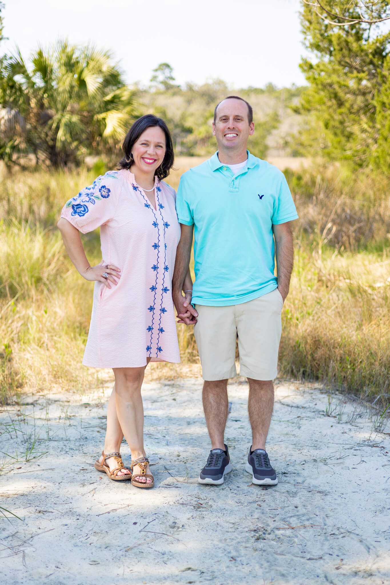 A happy couple stand holding hands in a pink dress and green shirt in a sandy park trail
