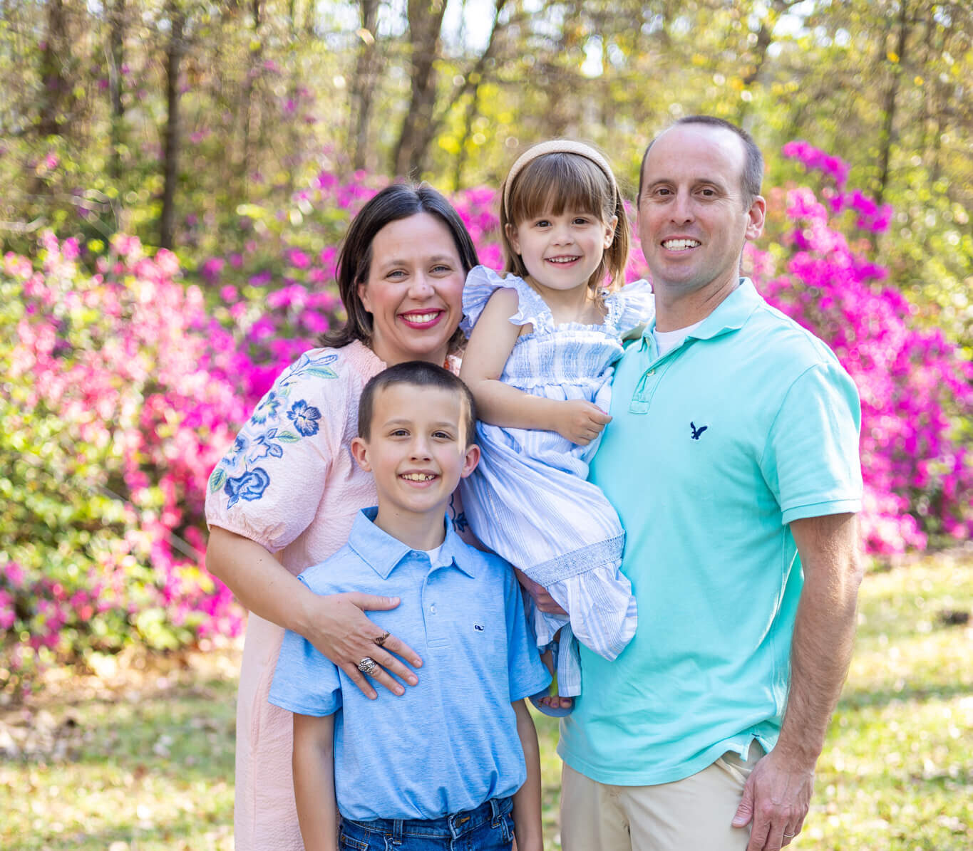 A dad holds his toddler daughter while standing with his wife and young son in a park lawn with blooming flowers after staying at a romantic bed and breakfast in savannah ga