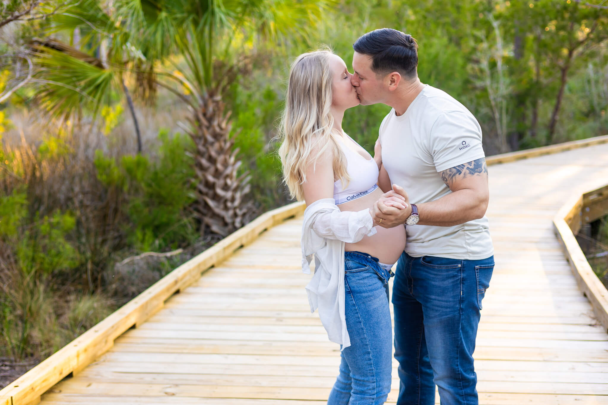 An expectant couple dances and kisses on a boardwalk at sunset in jeans and white shirts