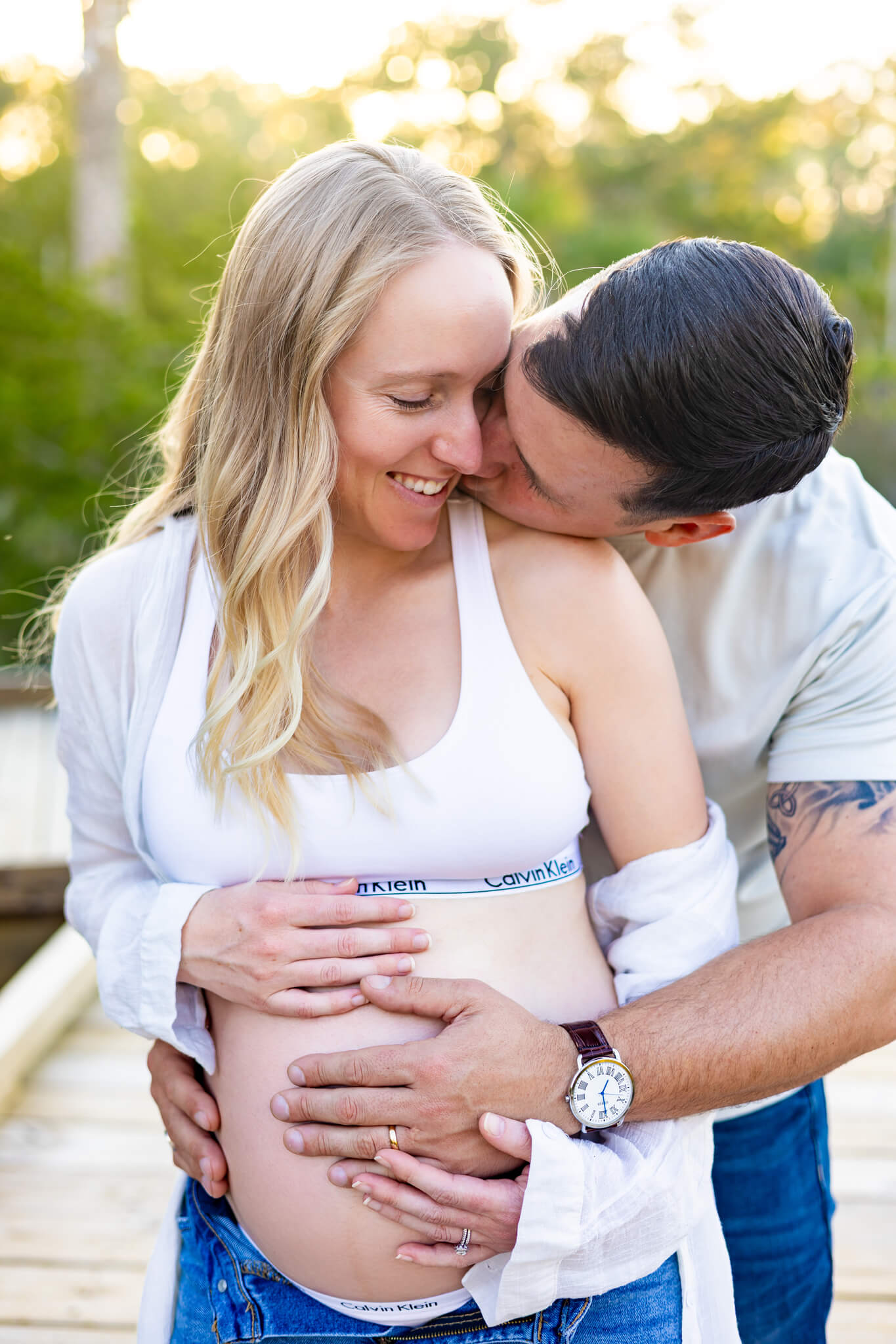 A happy pregnant couple snuggles and kisses with hands on the exposed bump on a boardwalk at sunset after finding romantic hotels in savannah, ga
