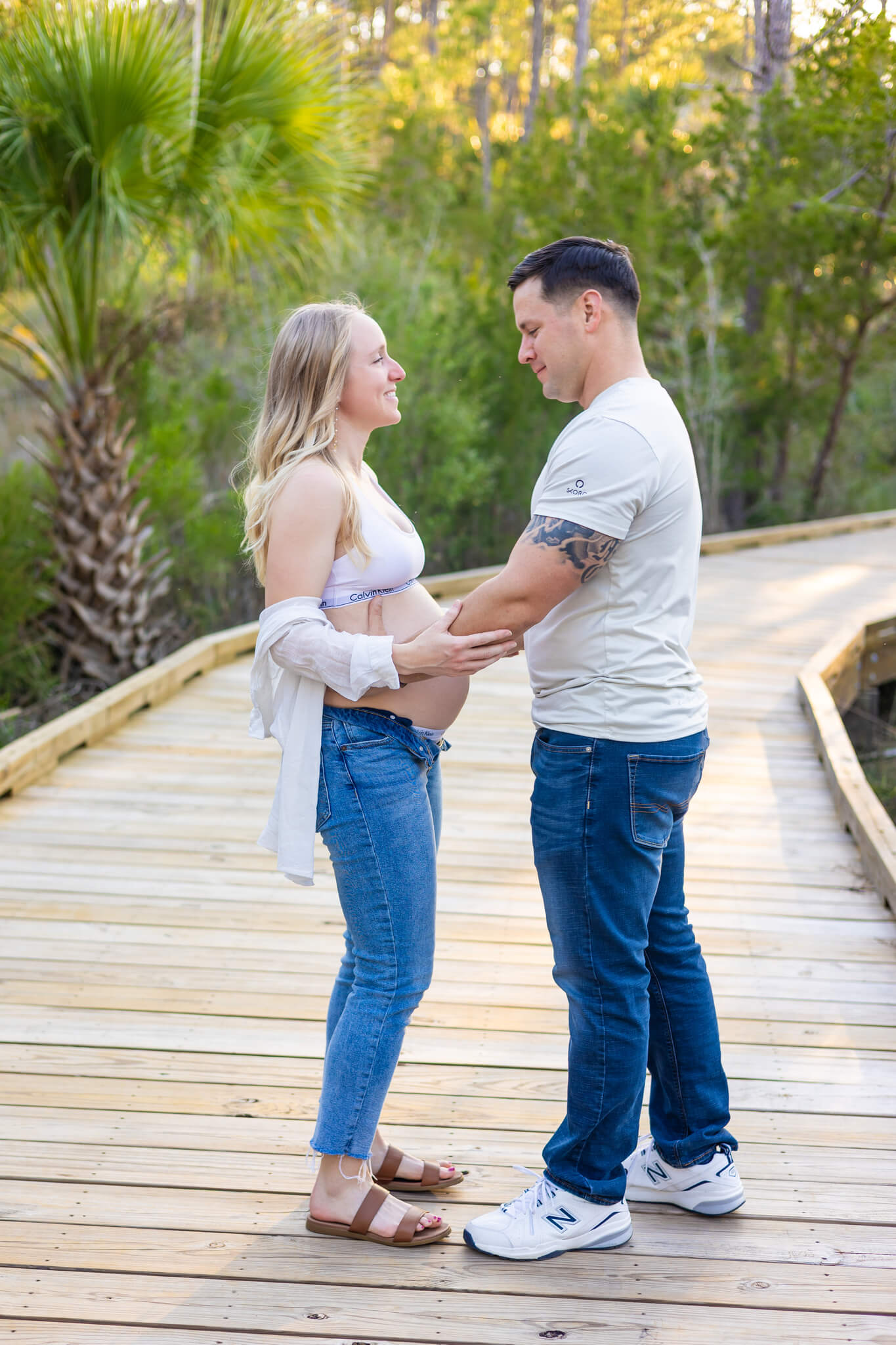 A happy expecting couple stands on a boardwalk at sunset admiring the bump in jeans and white shirt while staying at romantic hotels in savannah, ga