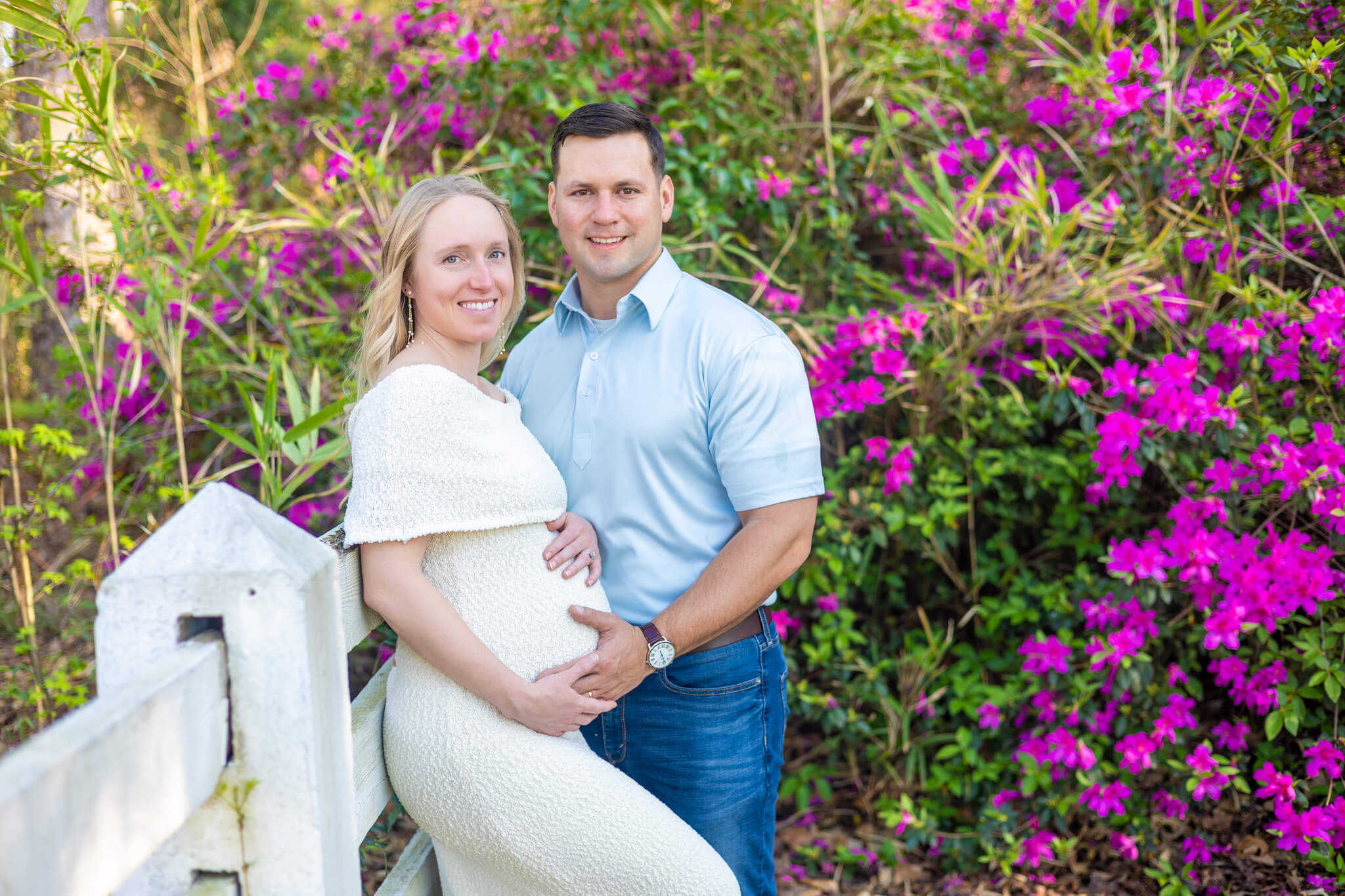 A smiling mom to be leans against a white fence in an azalea garden in a white maternity gown with husband in blue