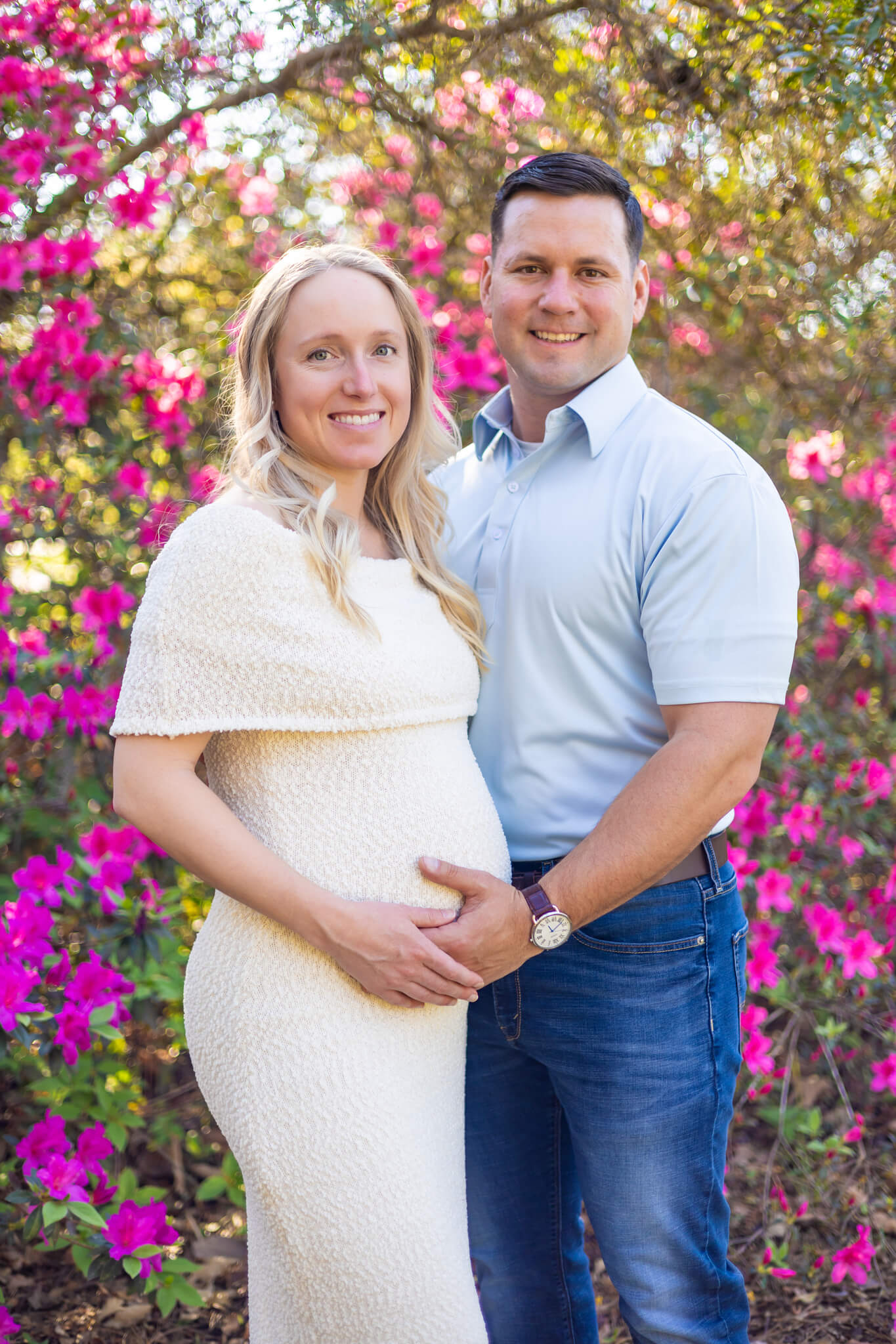 A smiling expectant couple standing among pink azaleas in a whtie maternity gown and white button down during a romantic weekend in savannah ga