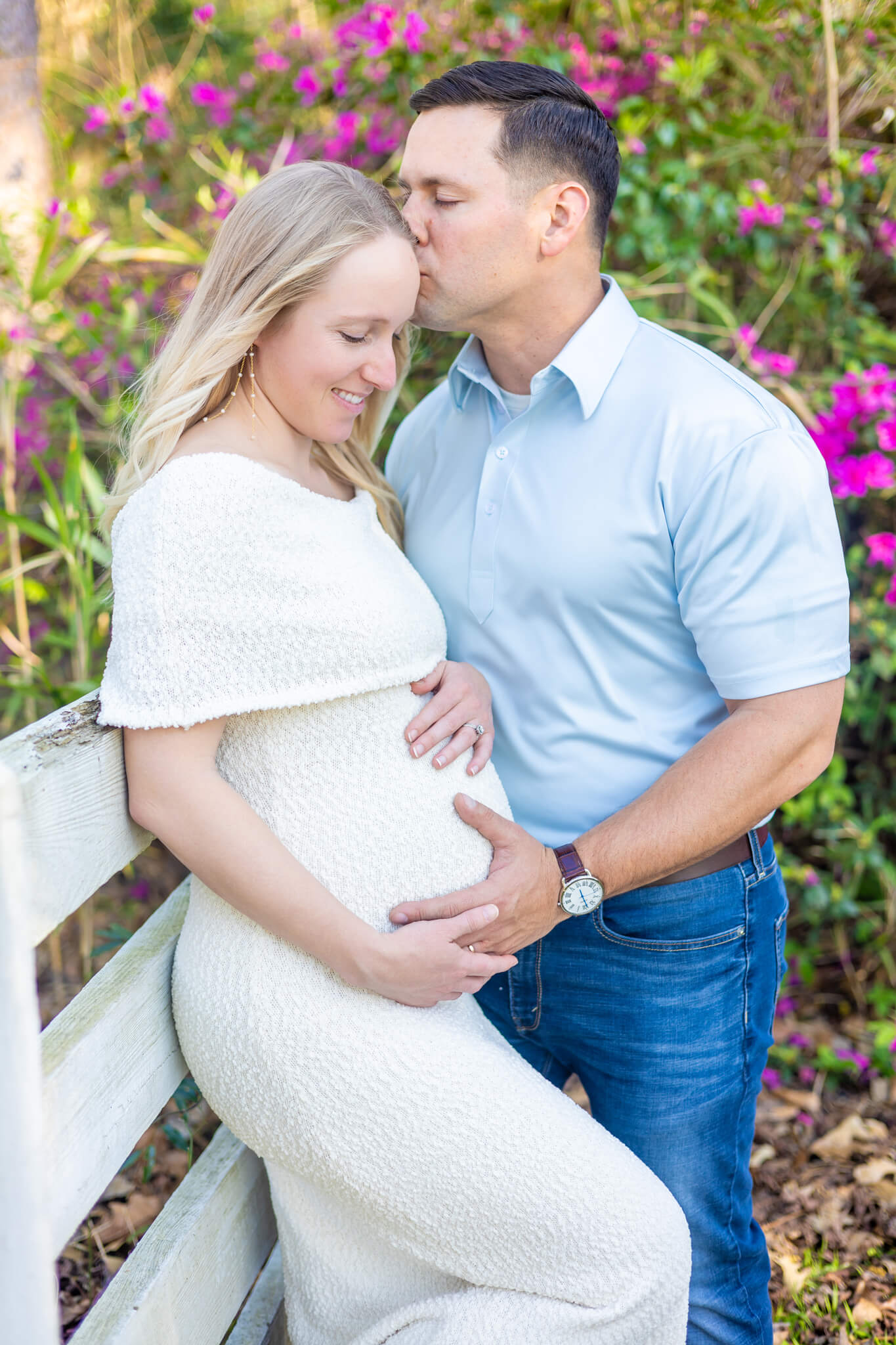 A happy pregnant couple lean on a rustic fence in a garden in white and blue with hands on bump during a romantic weekend in savannah ga