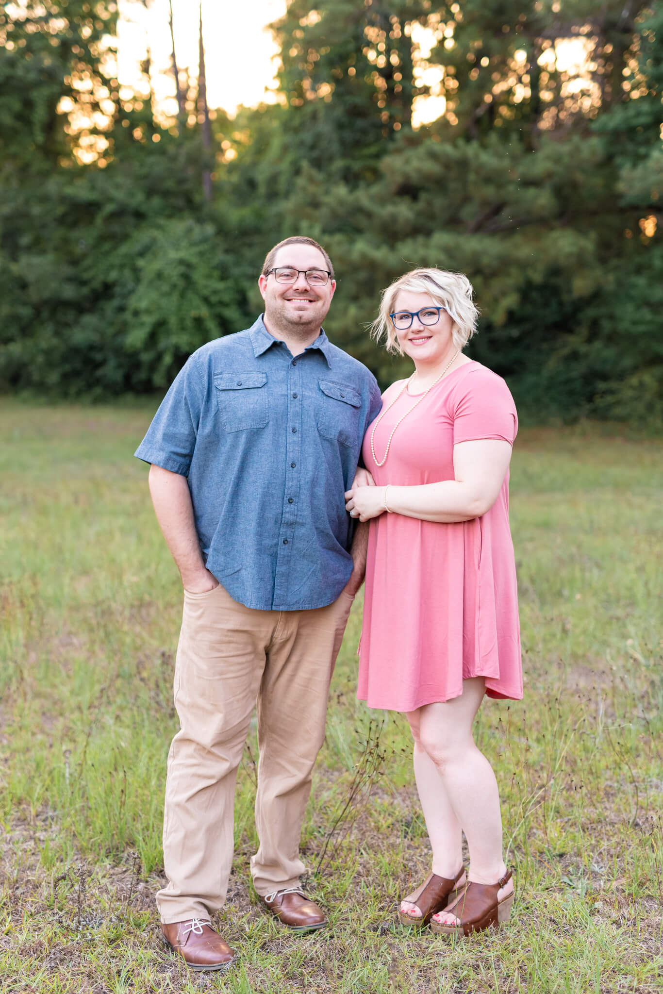 A happy couple in blue and pink stand smiling in a park lawn at sunset