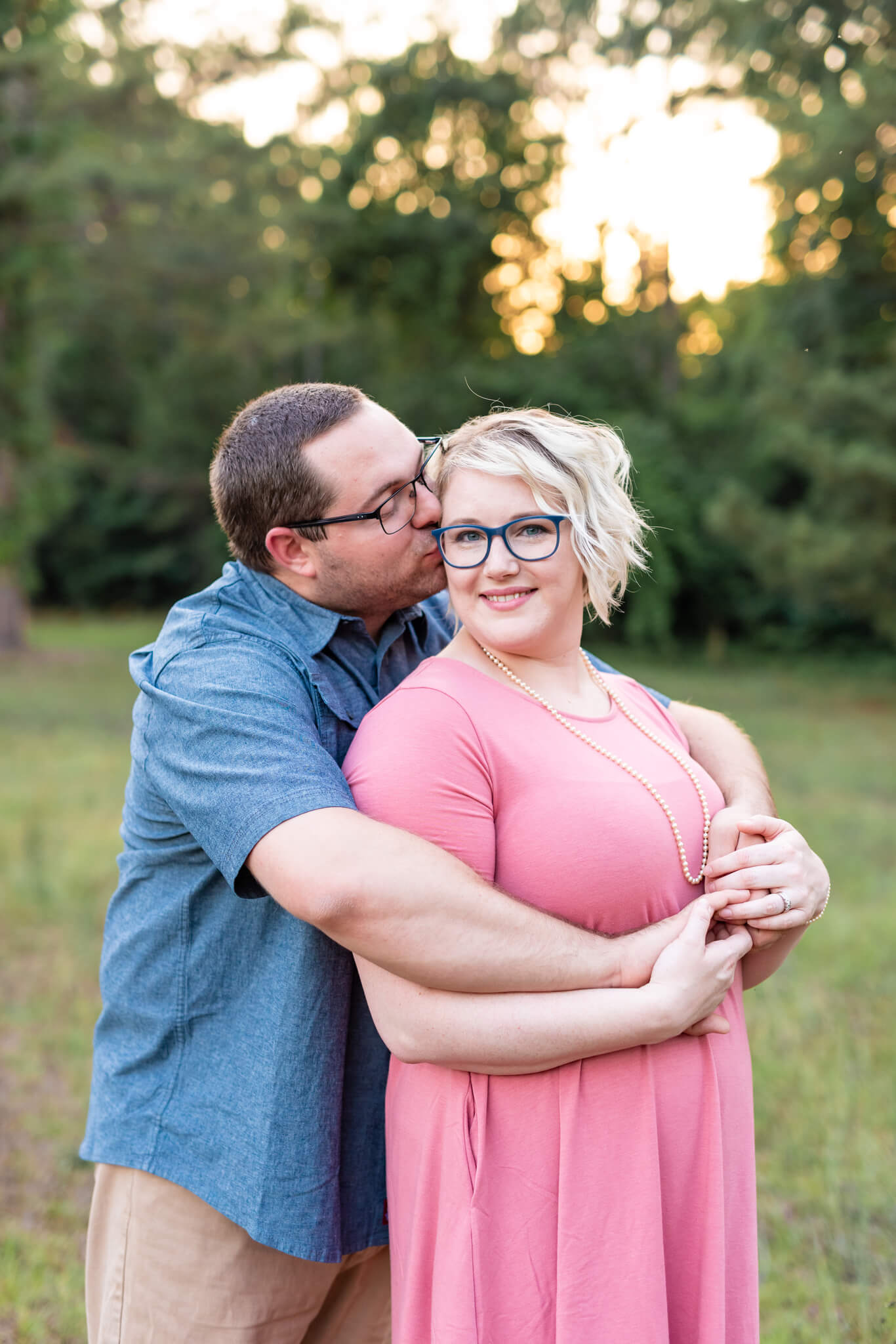 A man in blue kisses the cheek of his wife in a pink dress in a park lawn before heading to a sunset cruise in Savannah, GA