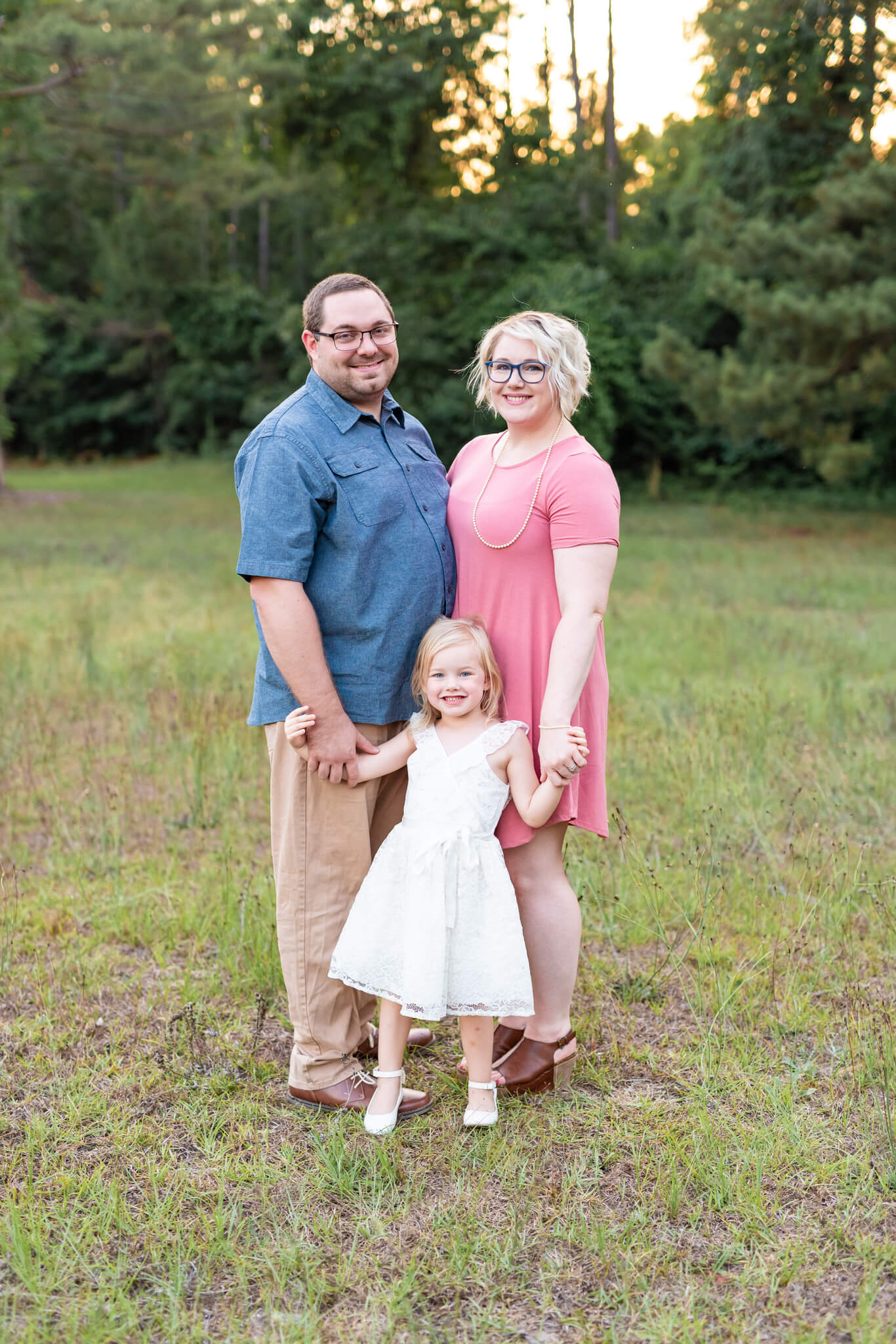 A mom and dad in blue and pink stand smiling with their toddler daughter in a white dress in a park field at sunset before enjoying a sunset cruise in Savannah, GA